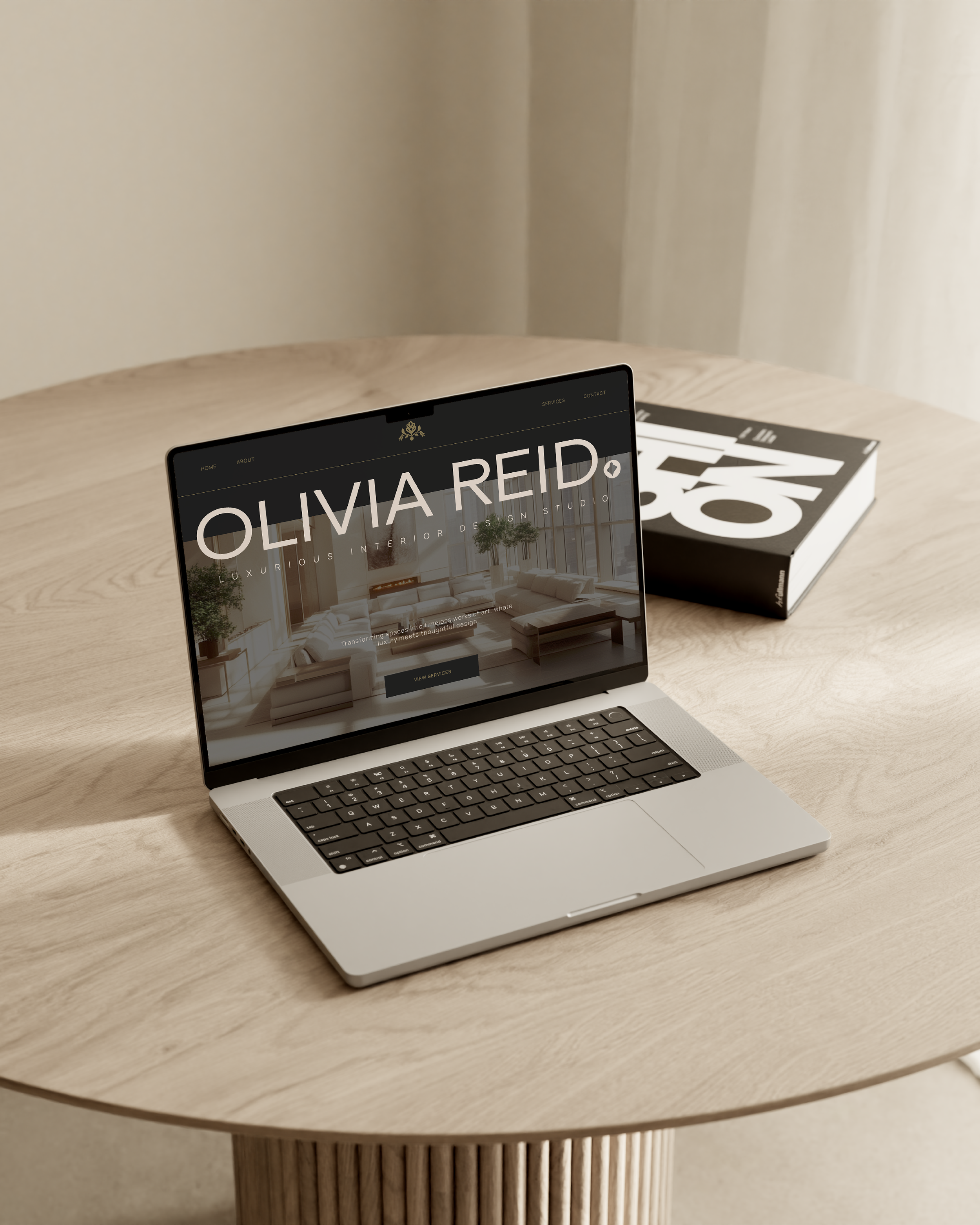 Open laptop on a wooden table displaying a interior design website with the name Olivia Reid, alongside a large black and white book.