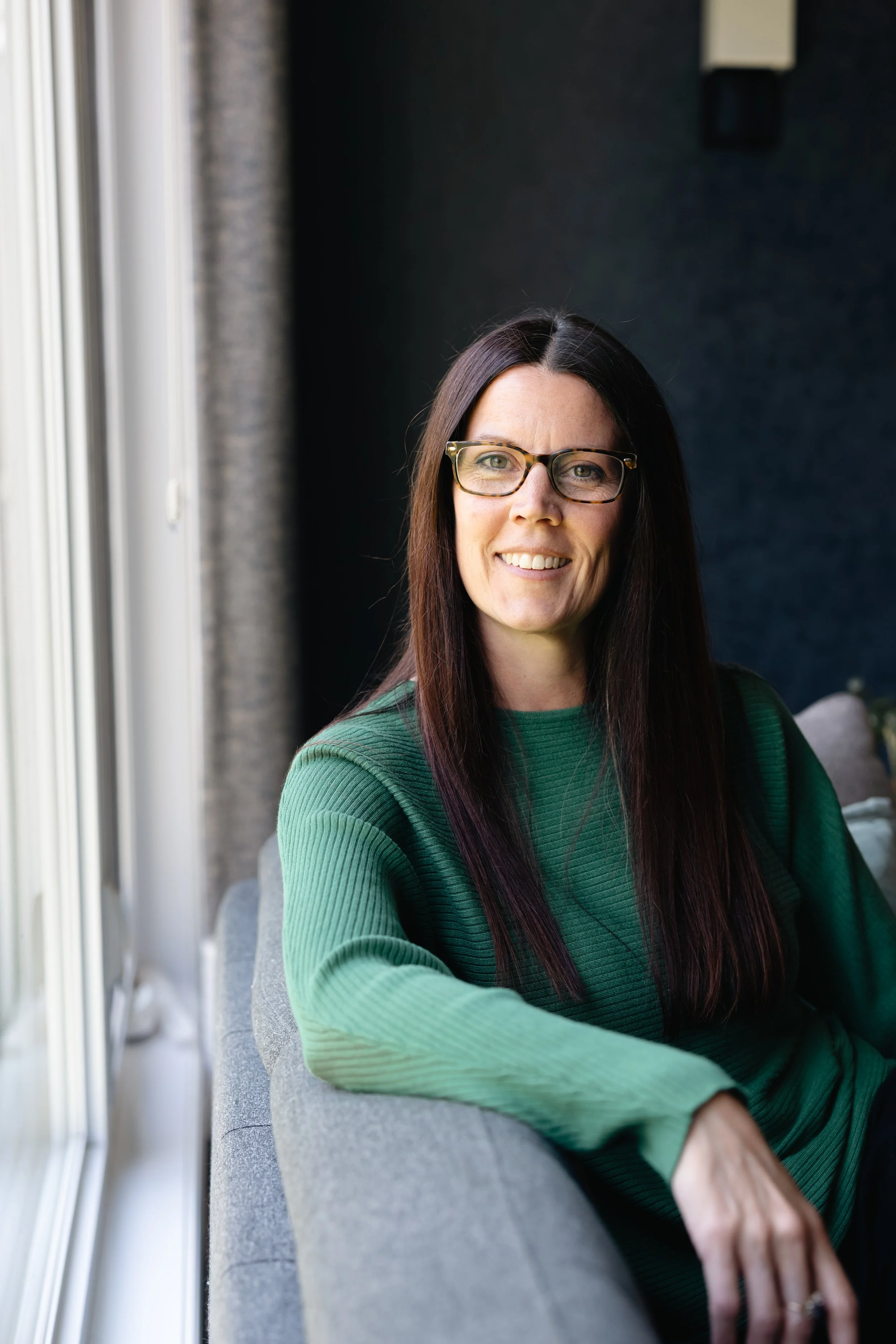 A woman wearing glasses and a green sweater, sitting on a gray couch near a window, smiling at the camera.