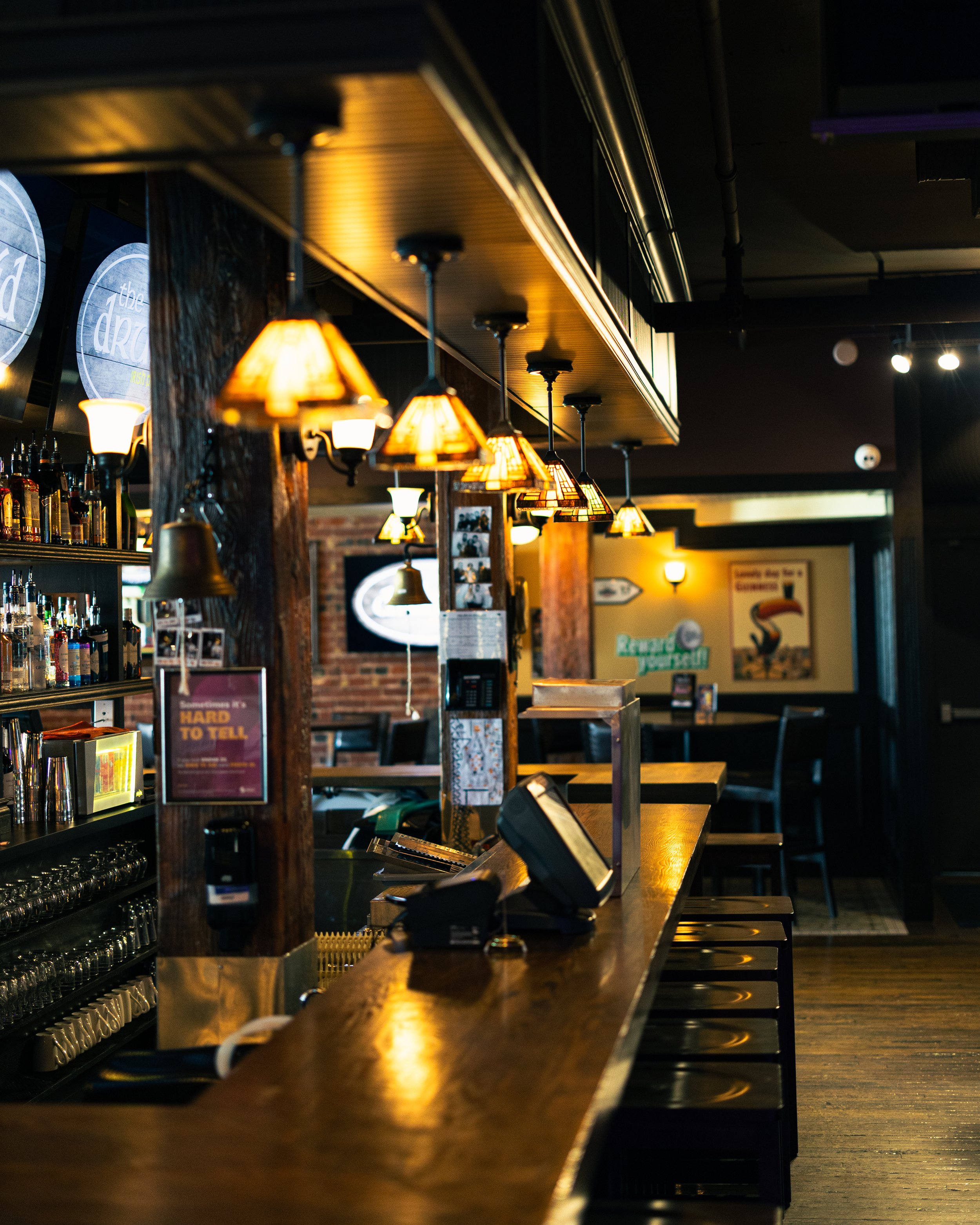 Interior of a bar or restaurant featuring hanging pendant lights with warm yellow glow, a long wooden counter with bar stools, shelves with bottles behind the counter, and a wall with promotional posters and decorations in the background.