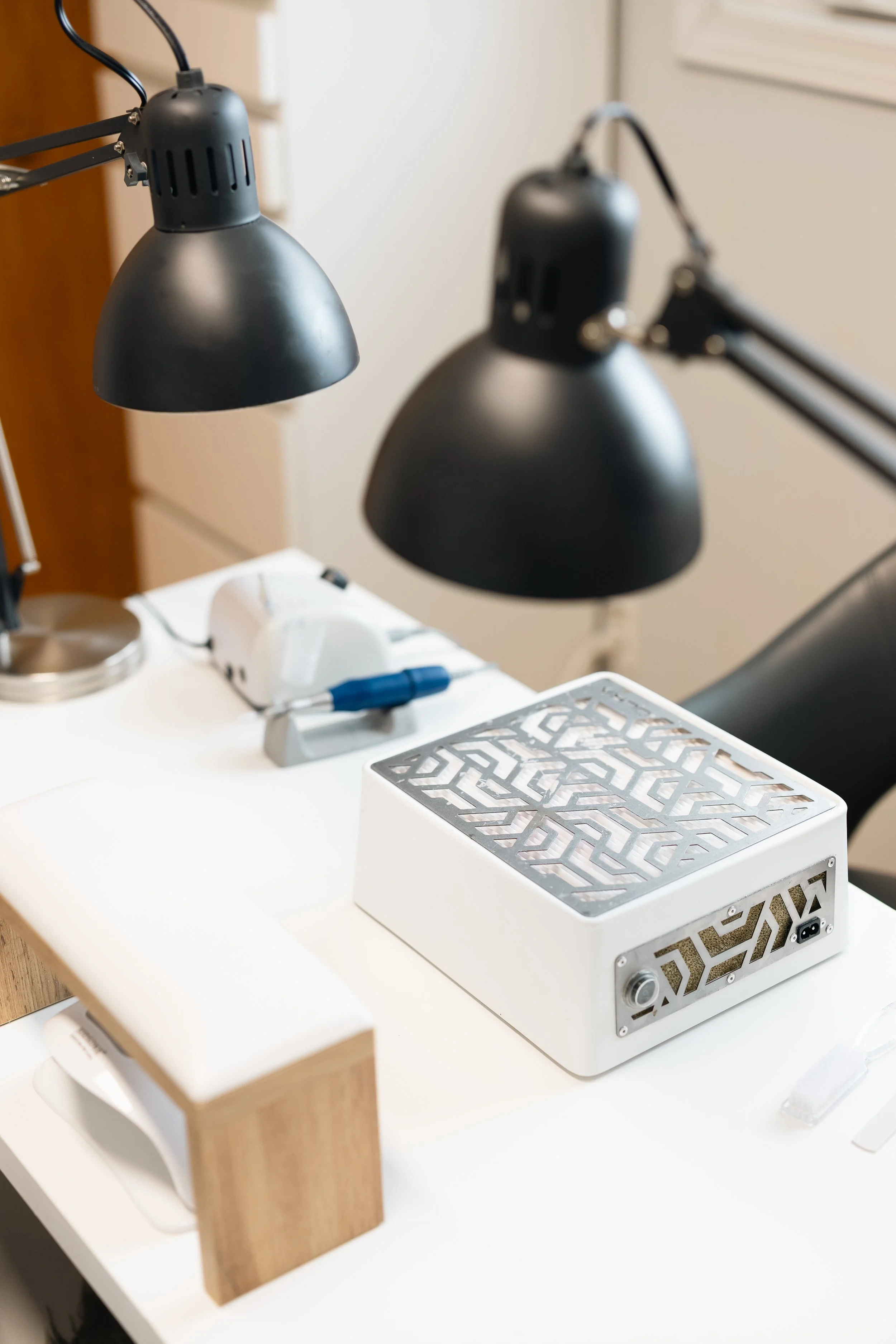 A white desk with two adjustable black lamps, a small wooden block, a device with a metallic patterned cover, and various small electronic items.
