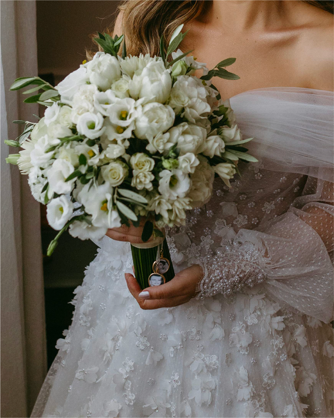 Close-up of a bride holding a bouquet of white flowers, wearing a white wedding dress with floral and bead embellishments, and a sheer long sleeve with lace and pearl details.