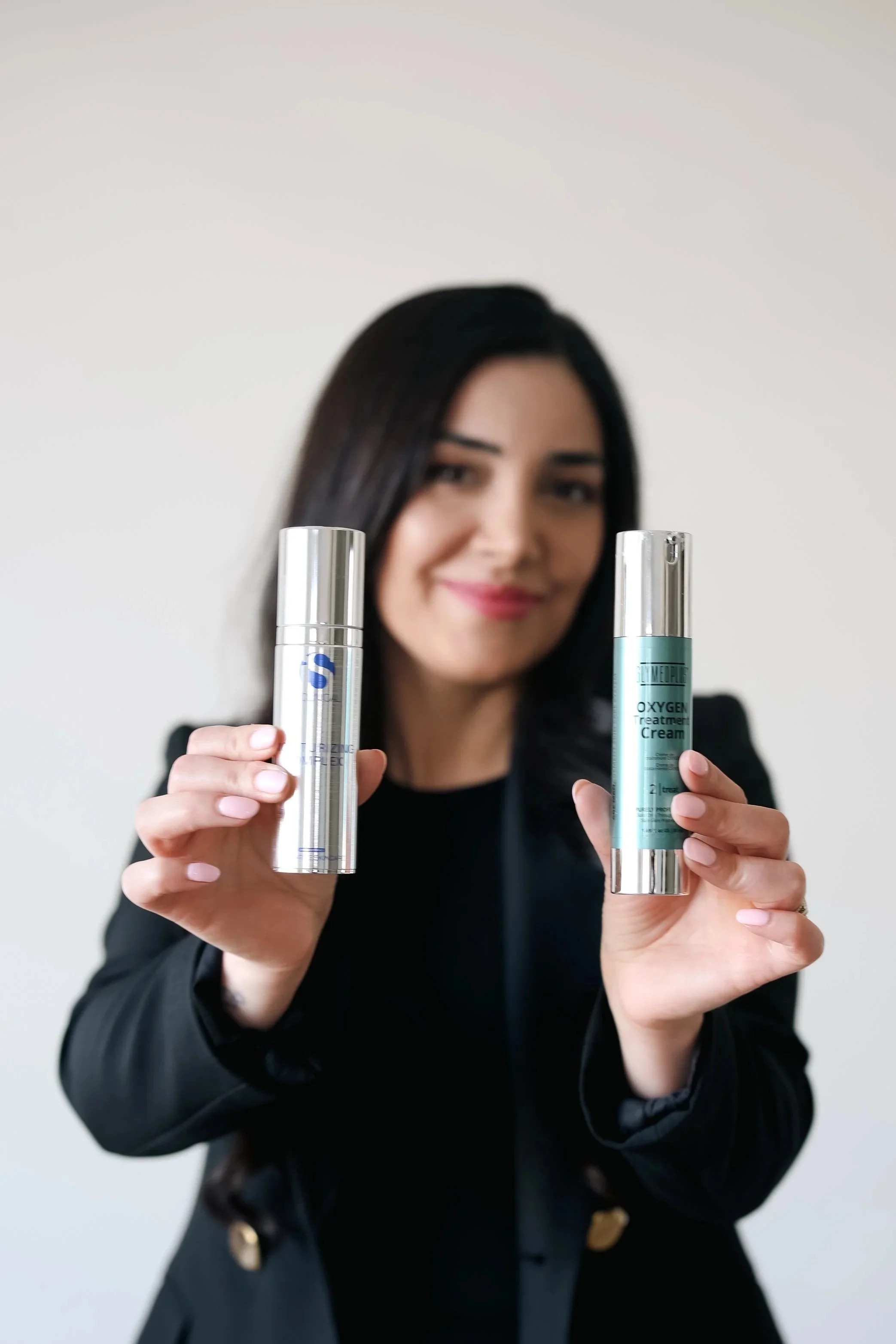 A woman holding skincare products, a blue oxygen treatment cream and a silver pump bottle, smiling slightly.