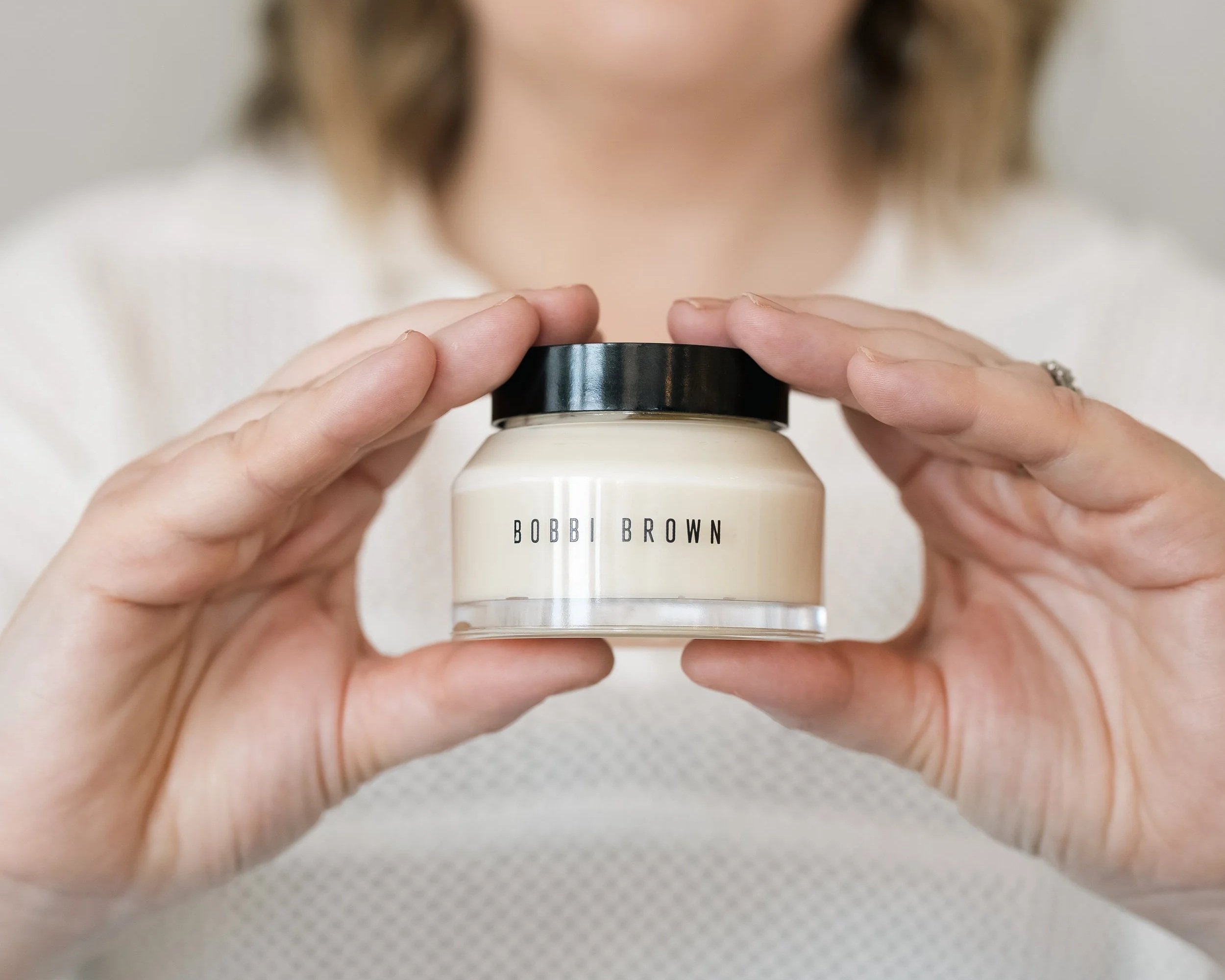 Person holding a jar of Bobbi Brown cream or skincare product with both hands, close-up view.