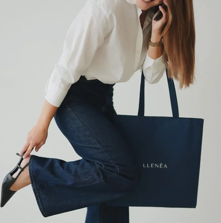 A woman in business casual attire, wearing a white blouse and dark blue jeans, bent over slightly to adjust her black pointed-toe heels while holding a large navy tote bag with the word 'LLENÉA' on it, in front of a plain light background.