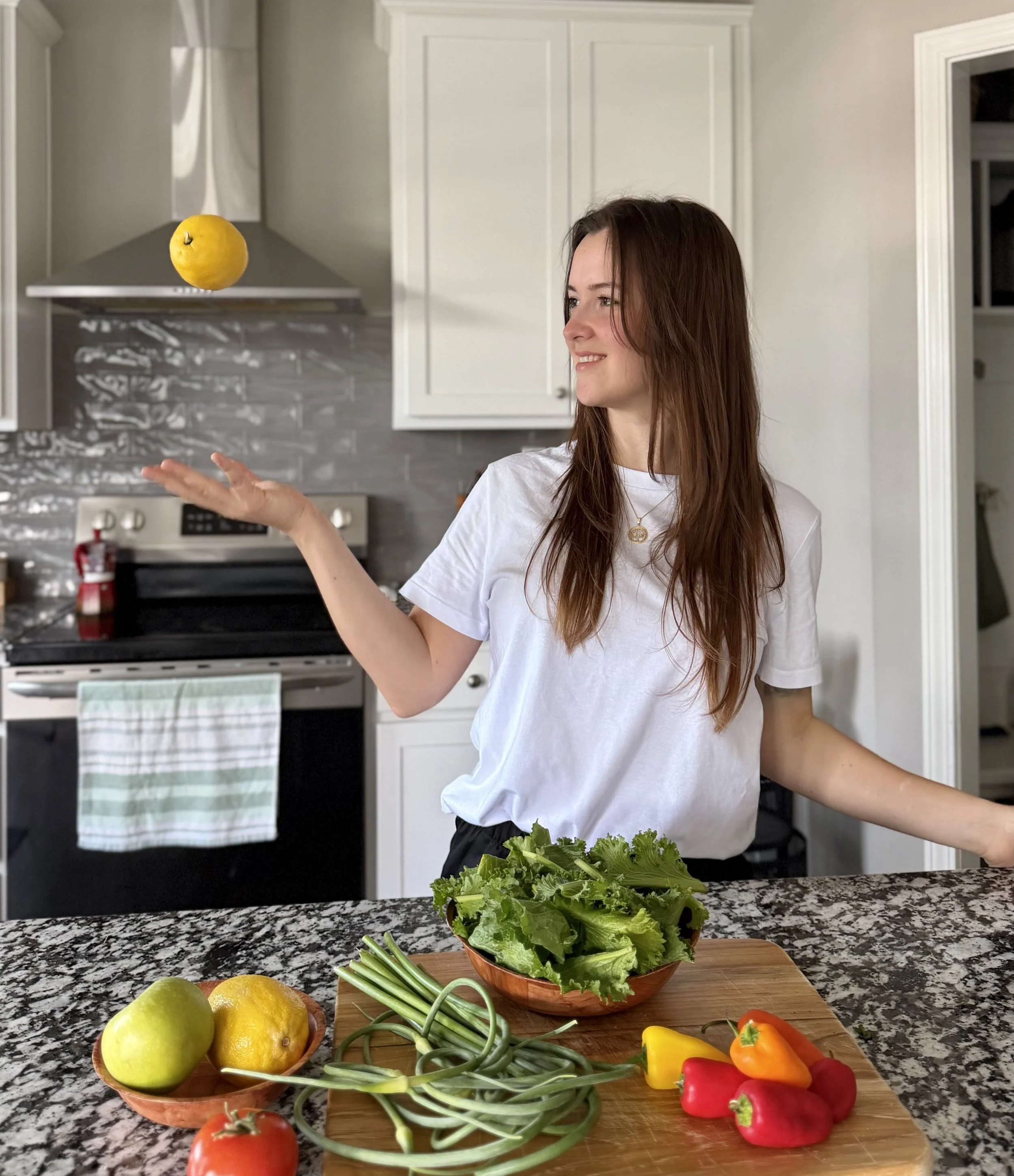 A woman with long brown hair wearing a white T-shirt is standing in a modern kitchen, smiling, and tossing a yellow lemon into the air. On a granite kitchen counter, there is a wooden cutting board with fresh vegetables including green leafy lettuce, green onions, orange, red, and yellow bell peppers, a tomato, and apples. A bowl of lemons and apples is also visible on the counter.