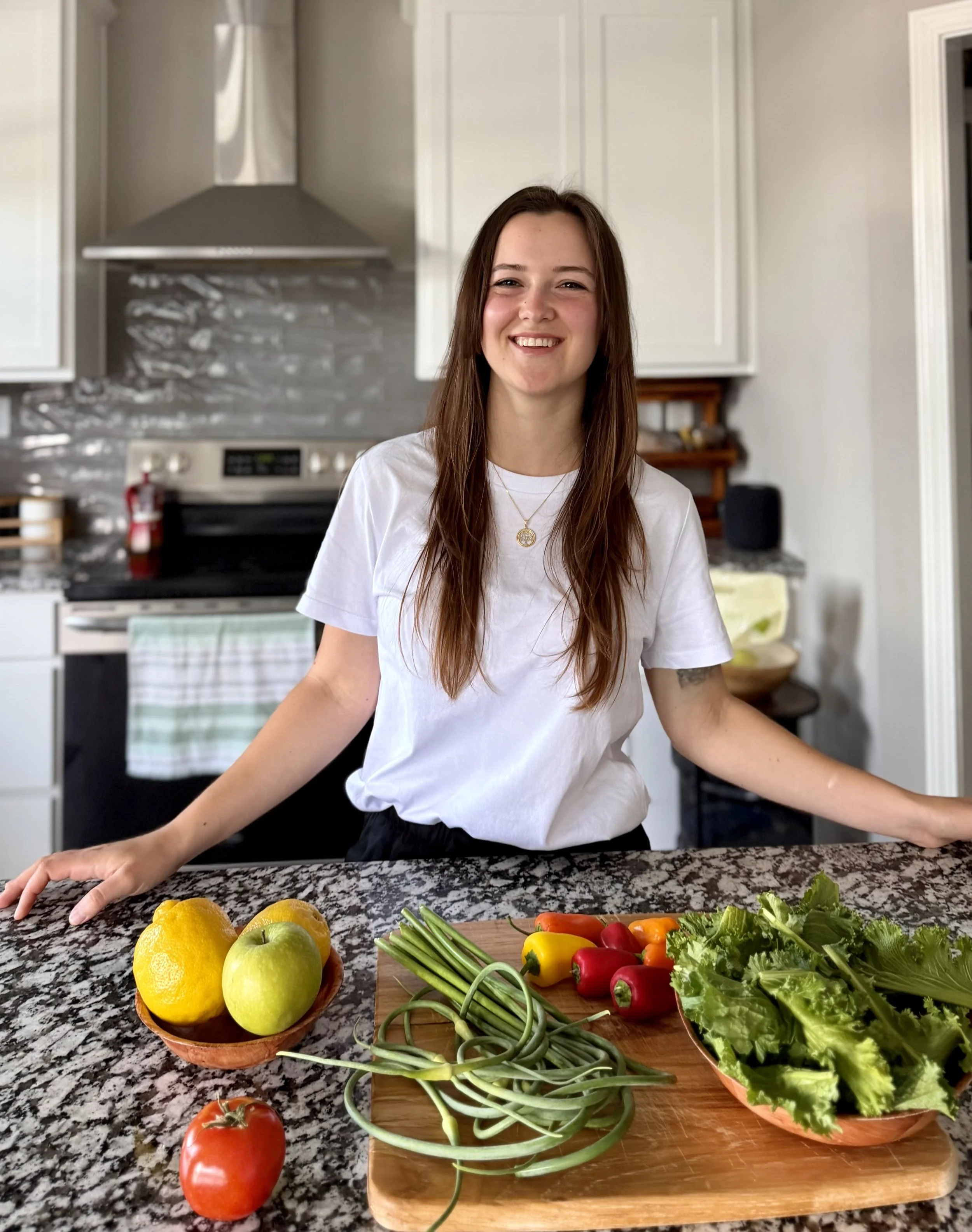 Woman smiling in a kitchen with fresh vegetables including lemons, green apples, tomatoes, lettuce, and bell peppers on a granite countertop and wooden cutting board.