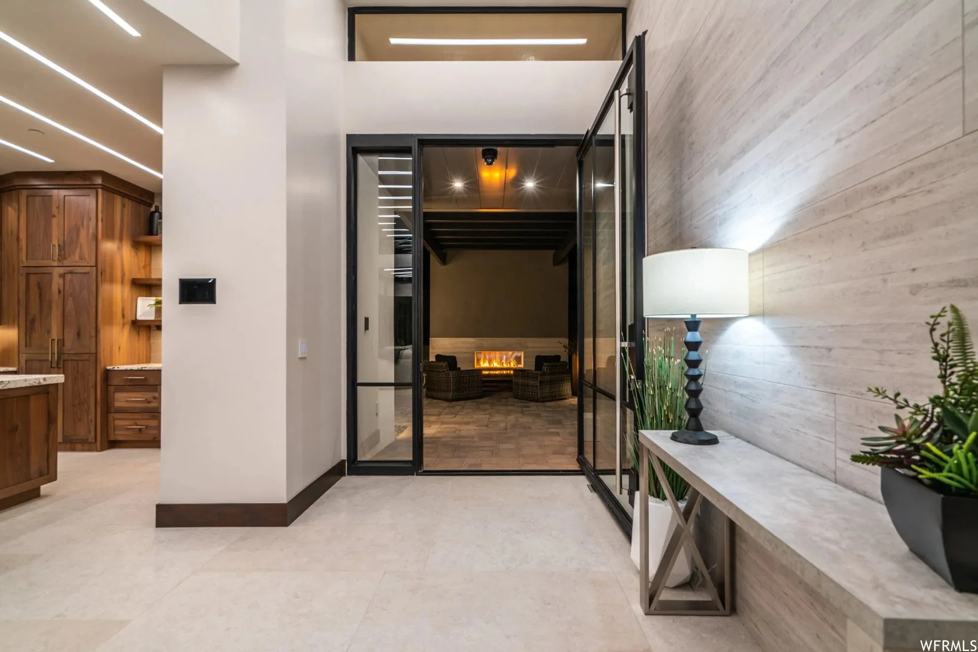 Modern interior with a hallway featuring a white wall, a marble table with a lamp and plants, and a glass sliding door leading to a sitting area with chairs and a fireplace.