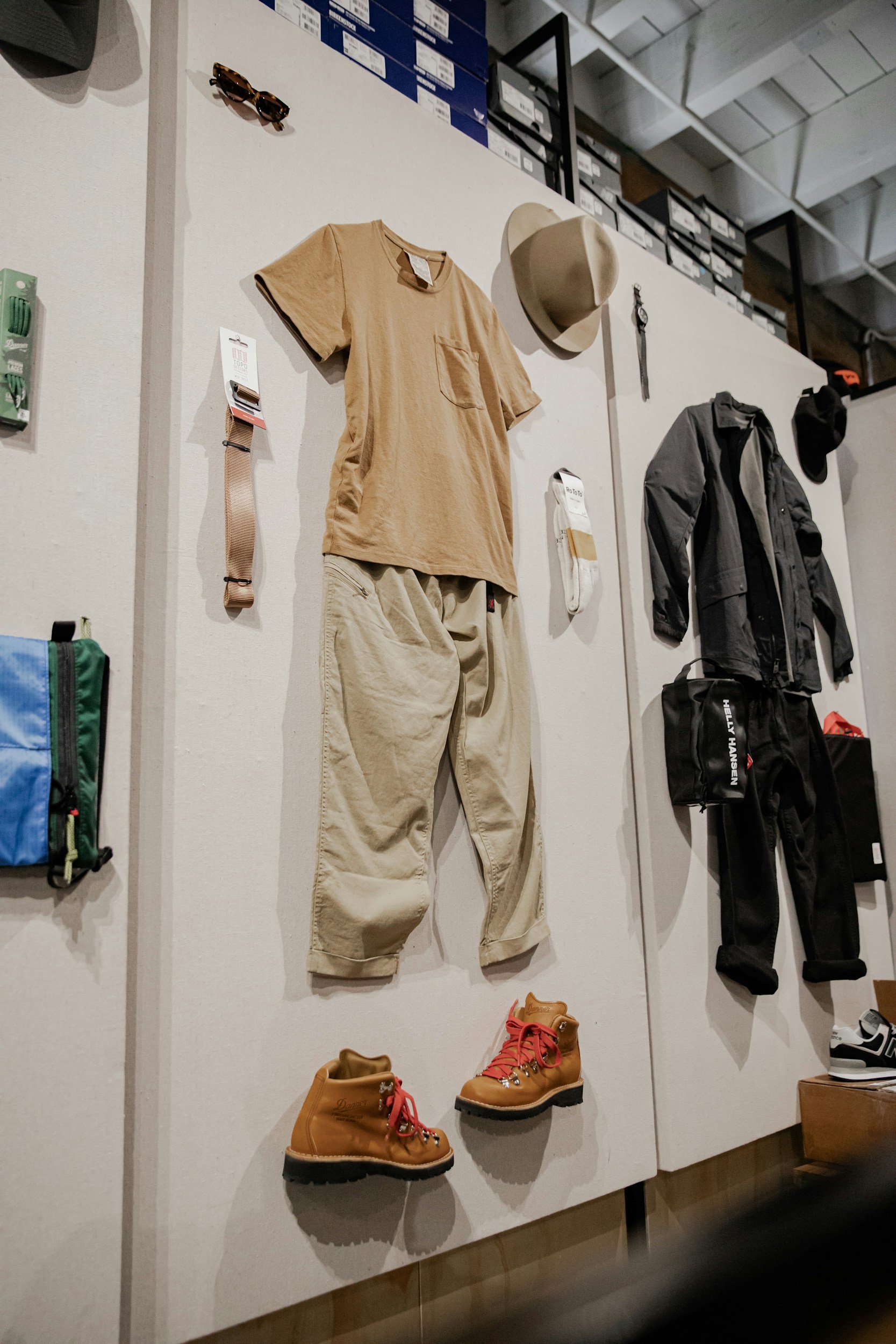 Display of outdoor apparel including a beige t-shirt, beige pants, brown hiking boots, a beige hat, and various accessories on a wall at a retail store.