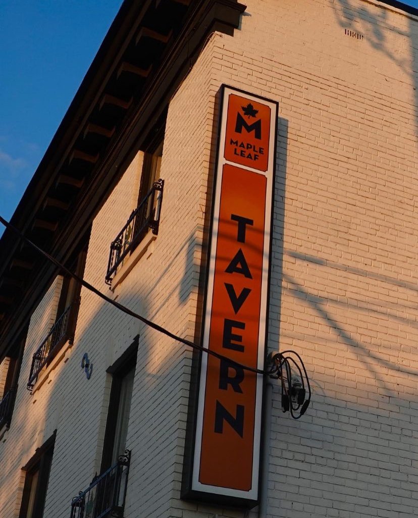 Vertical sign on a white brick building with a maple leaf logo at the top, reading 'MAPLE LEAF' and 'TAVERN' in bold black letters on an orange background.