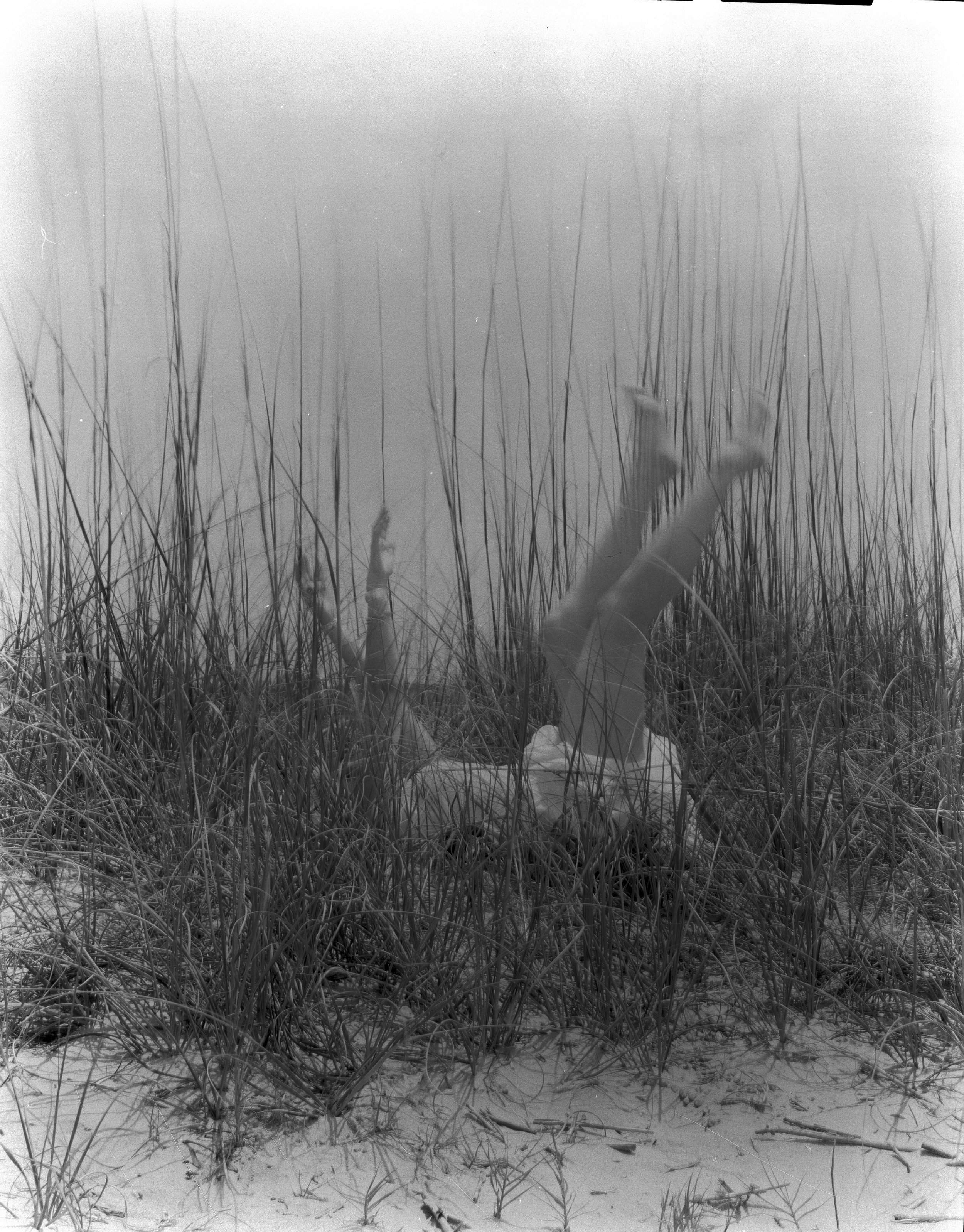 Person laying on the sand in a grassy area, with legs raised and feet in the air, viewed through tall grass.