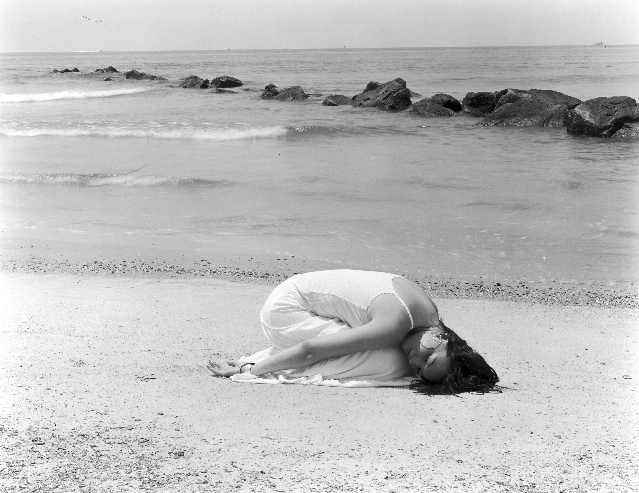 A woman wrapped in a blanket lying on the beach, with a rocky jetty and the ocean in the background.