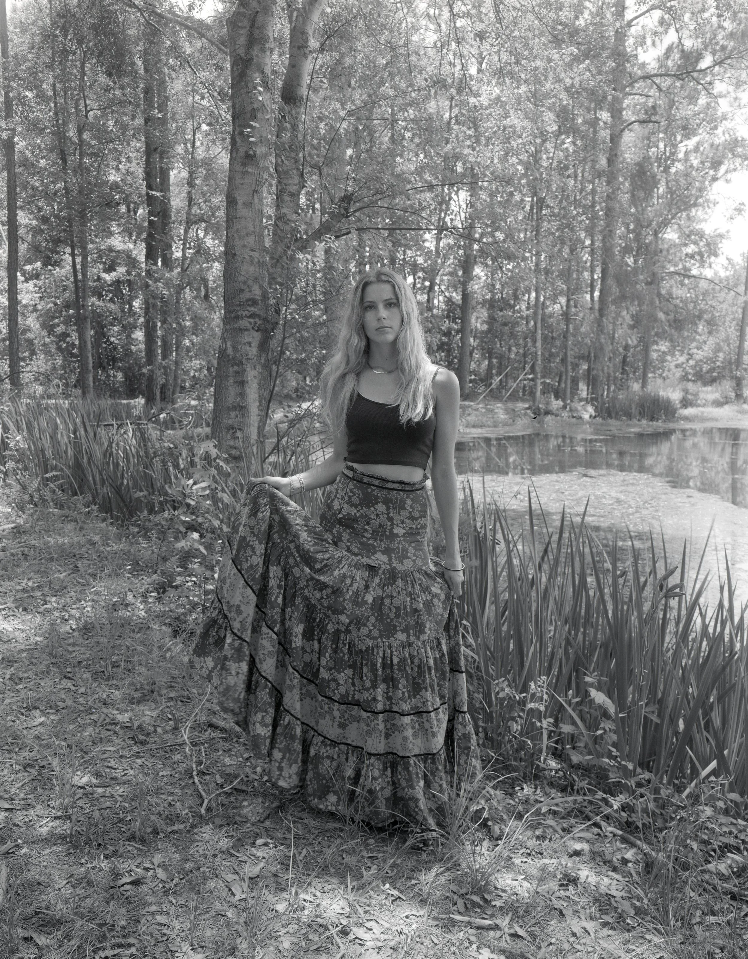 A young woman with long wavy hair standing outdoors by a pond, holding the edge of her long patterned skirt. She's wearing a black sleeveless top and appears to be in a wooded area.