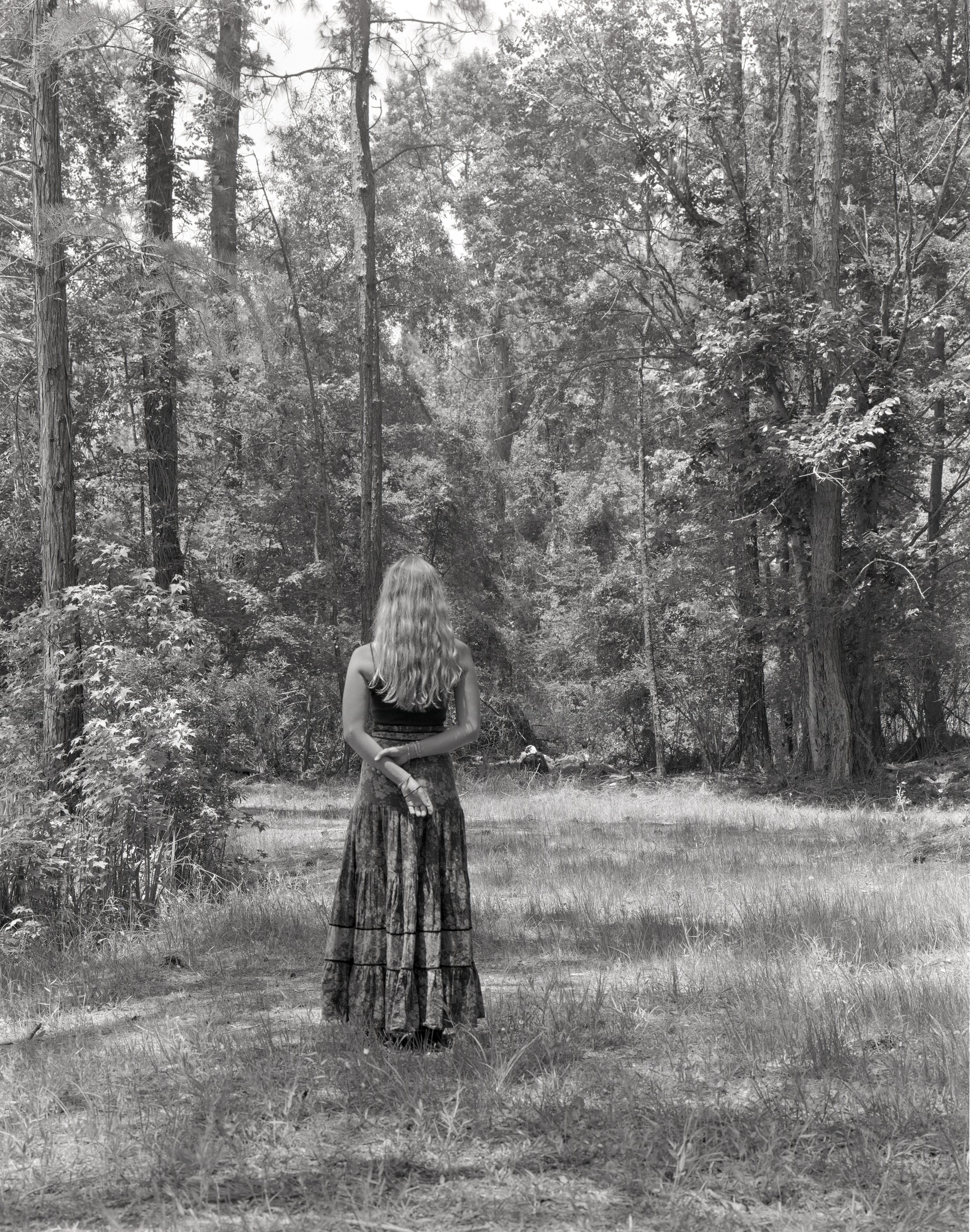 A woman with long, wavy hair stands with her back to the camera in a forest, surrounded by tall trees and grass.