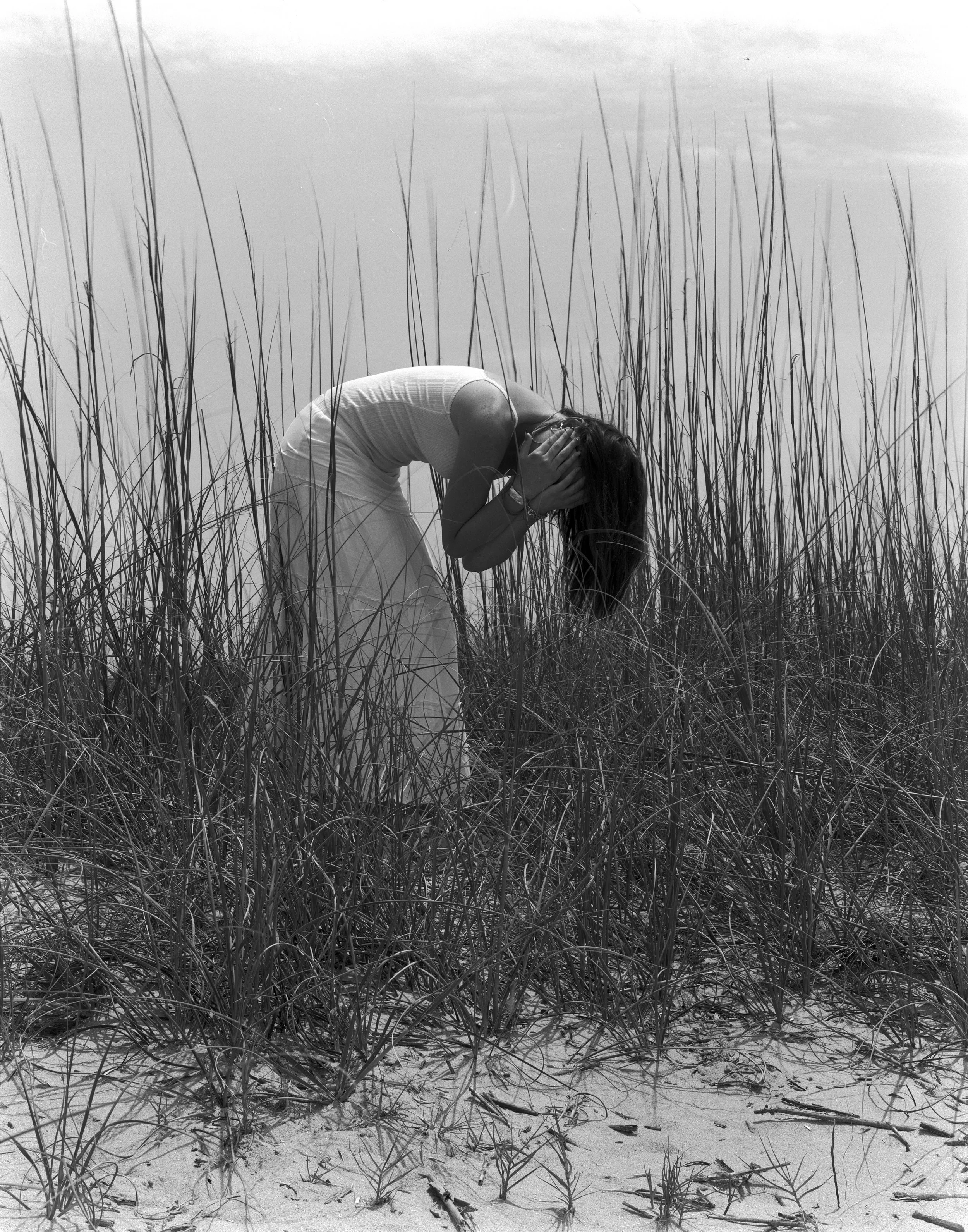 A woman in a white dress holding her head and bent over in a sandy, grassy outdoor setting.