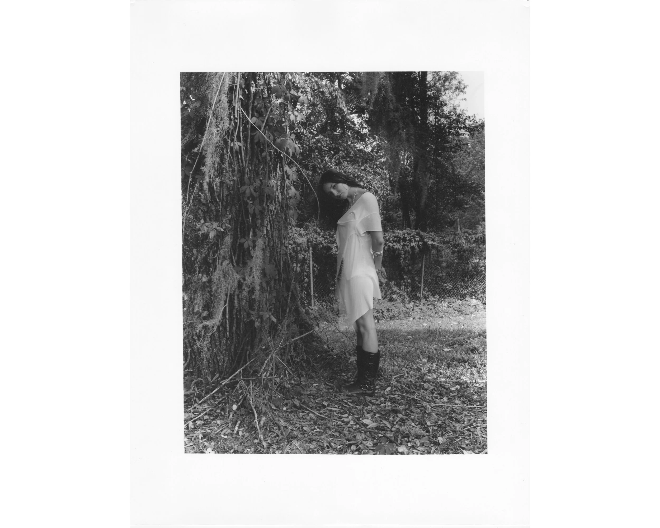 A woman wearing a light-colored dress and dark boots standing outdoors near a large tree with hanging moss, in a natural, wooded area.