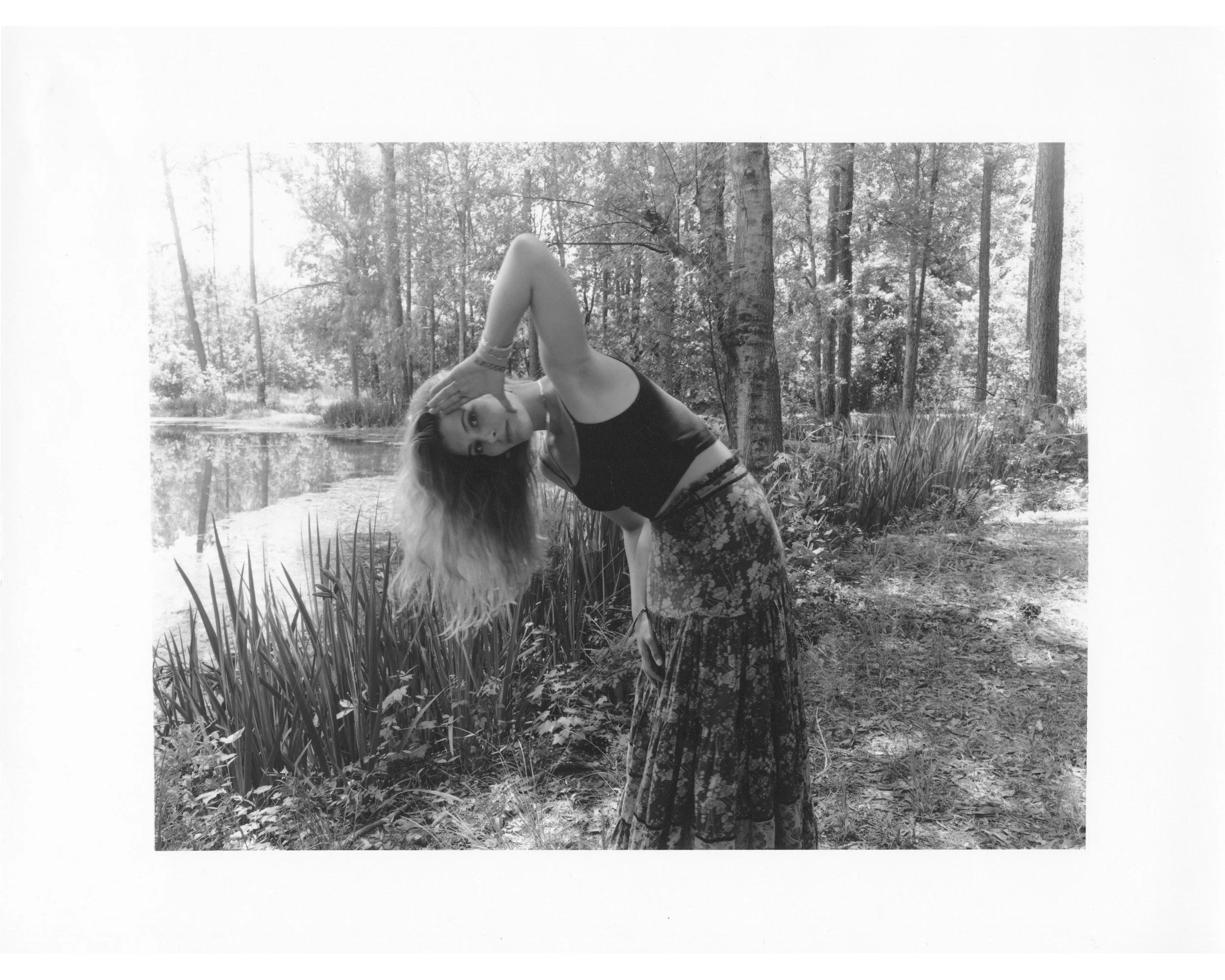 A woman with long wavy hair, wearing a black tank top and a long floral skirt, stands outdoors near a pond with trees and grass in the background. She is bending sideways with one arm behind her head and the other hand touching her knee, looking at t