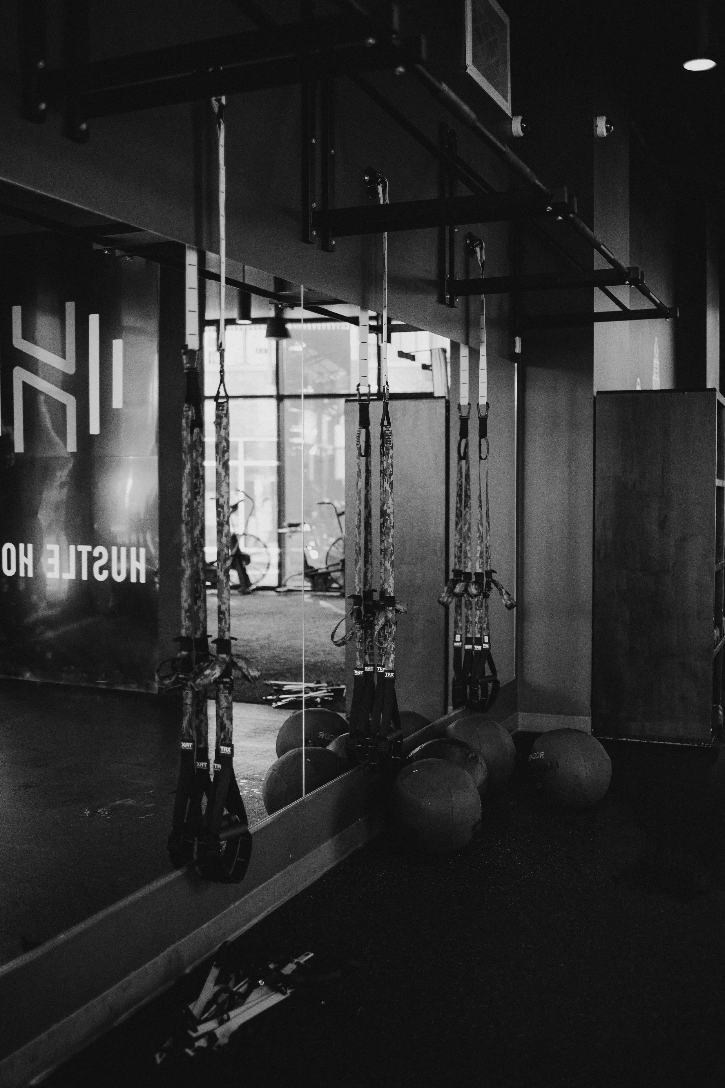 Interior view of a fitness studio with suspension training straps hanging in front of a large mirror, medicine balls on the floor, and bicycles outside the window, all in black and white.