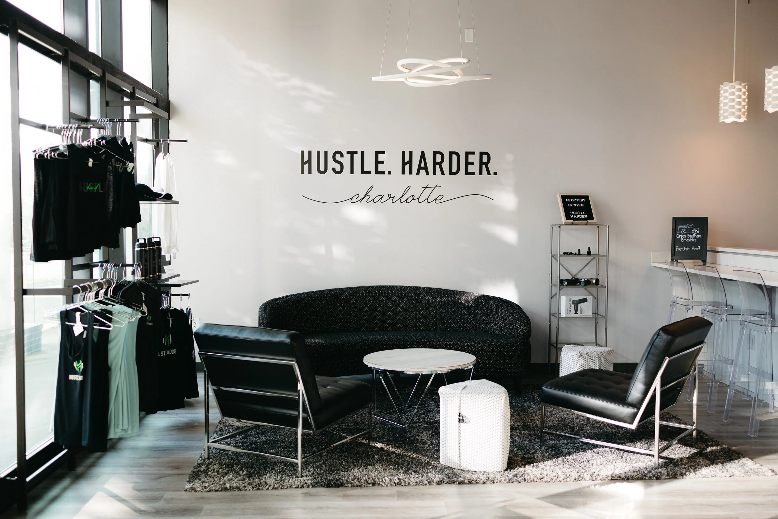 Modern lounge area with black sofa and chairs, round coffee table, and clothing display with fitness apparel, in a fitness or wellness center with motivational sign reading 'HUSTLE. HARDER. Charlotte' on the wall.