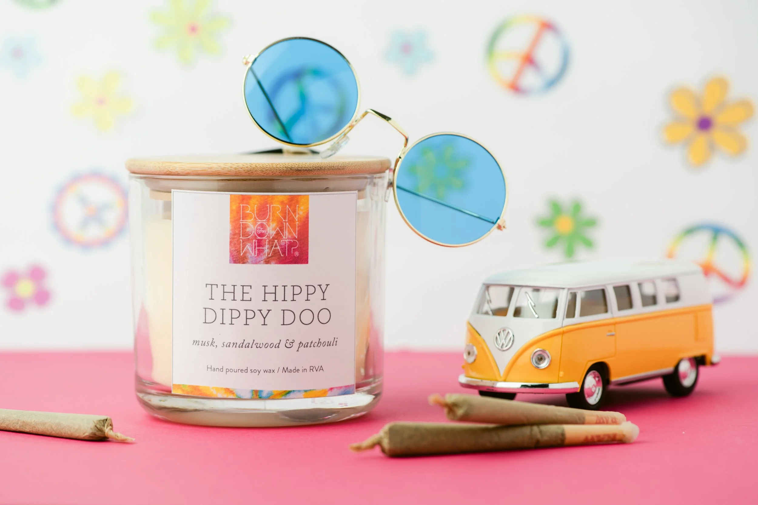 A scented candle labeled 'The Hippy Dippy Doo' with notes of musk, sandalwood, and patchouli, a miniature yellow and white Volkswagen bus toy, a pair of round sunglasses with blue lenses, and three rolled smudge sticks on a pink surface. Colorful pea