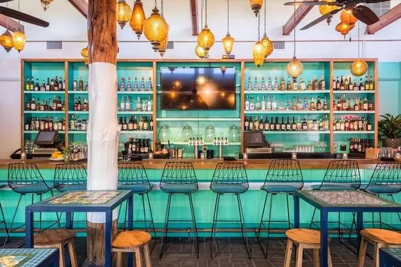 Colorful bar interior with a teal bar counter, shelves stocked with liquor bottles, and hanging lantern-style lights.