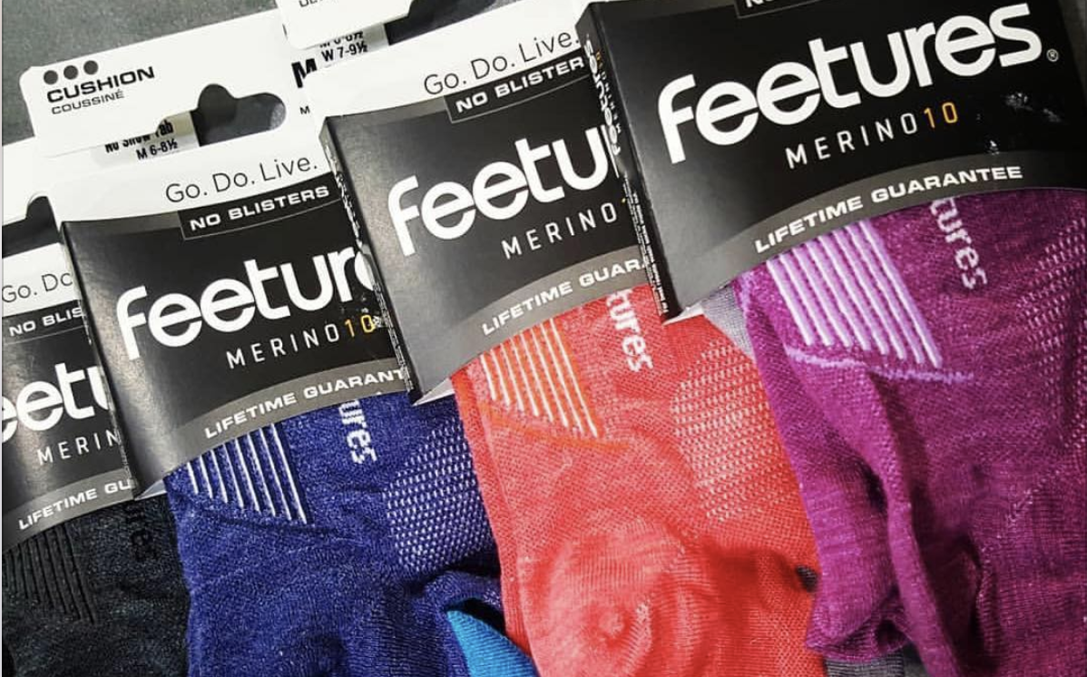 Multiple packages of Feetures Merino socks in various colors, including black, blue, red, and purple, with the packaging showing brands and product details.