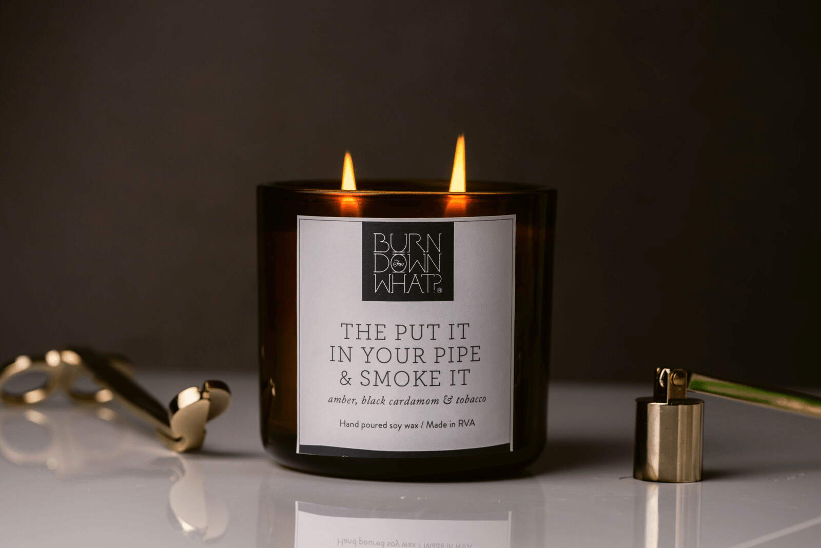A dark brown candle with two flames, labeled "Burn Down What?" and a humorous message about putting it in a pipe and smoking it, with scents of amber, black cardamom, and tobacco. The candle is placed on a white surface, with a gold item and a metall