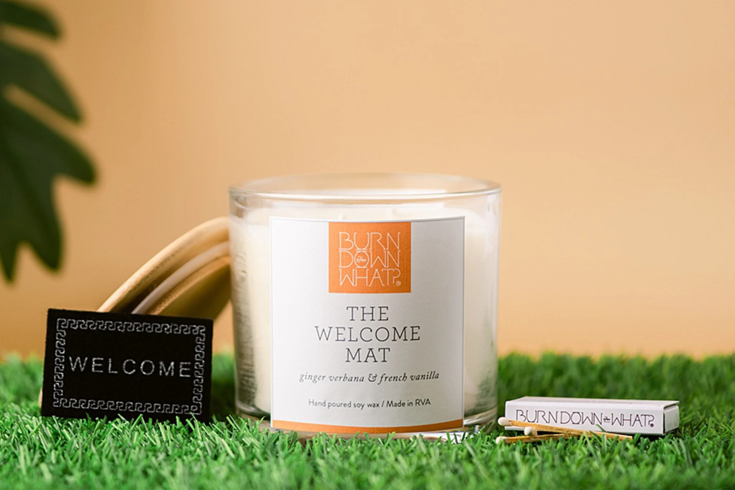 A white candle labeled "The Welcome Mat" with ginger verbena and French vanilla scent, surrounded by matches and a black welcome sign, on a patch of artificial grass with a beige background.