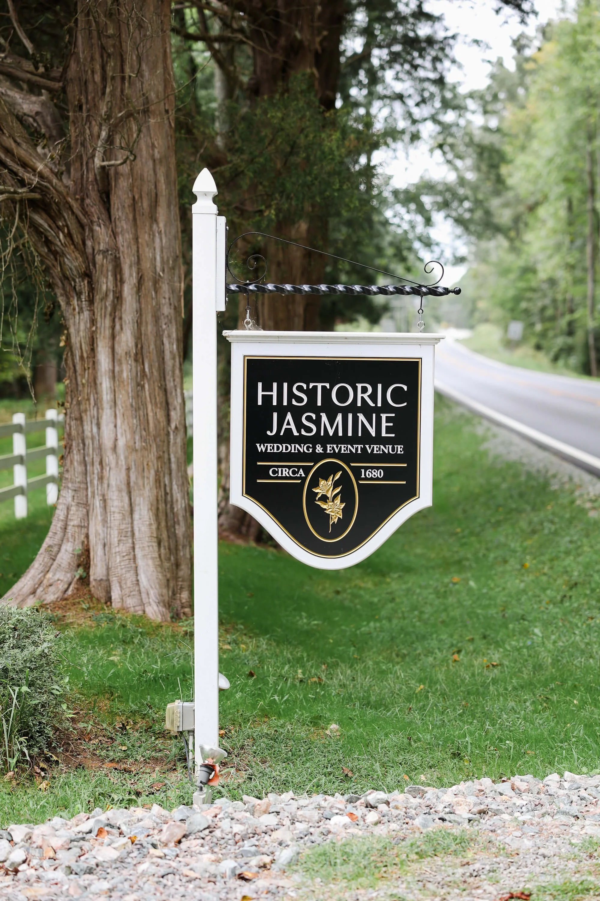 Signboard for Historic Jasmine, a wedding and event venue, mounted on a white post near a tree, with a rural road in the background.