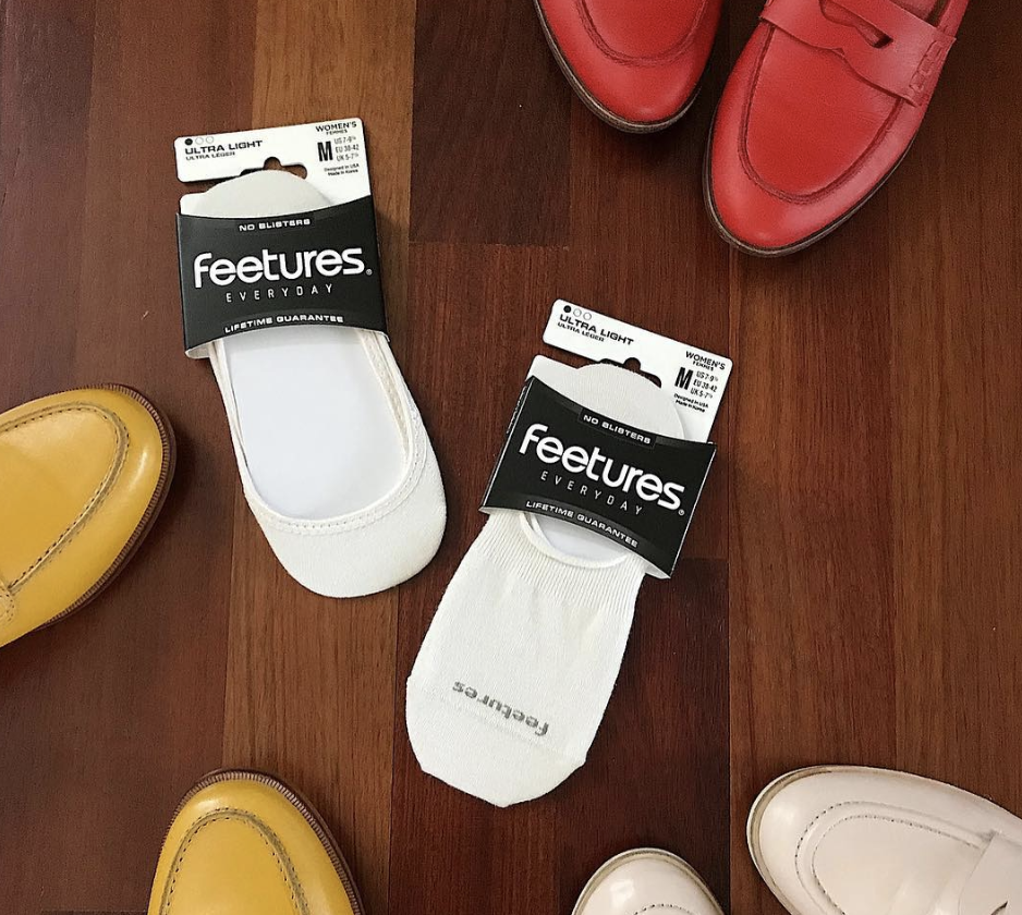 Two pairs of no-slip white footless socks with black labeling, one pair labeled for women, placed on a wooden floor. Surrounding the socks are shoes in yellow, red, and white colors.