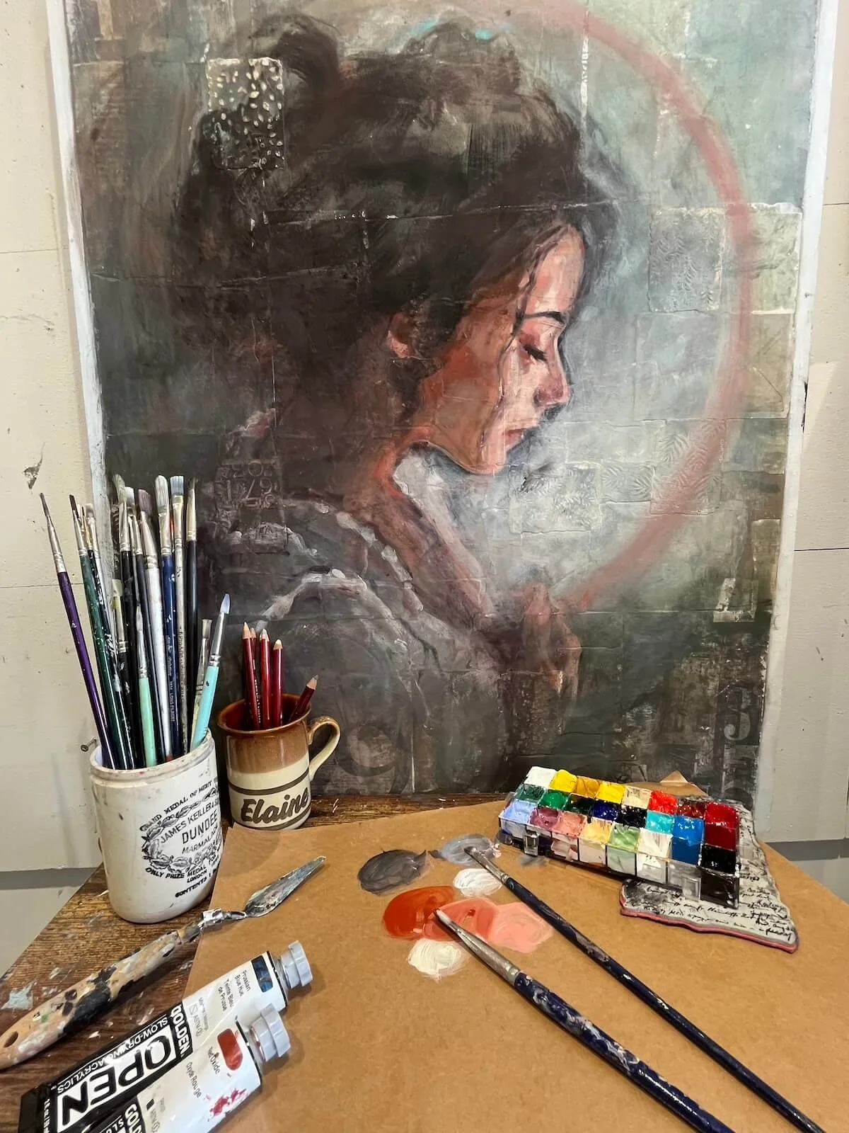 A painting of a woman with closed eyes, resting her chin on her hand, painted on a textured surface. In front of the painting is a table with various art supplies including paint tubes, brushes, a palette, and containers with colored pencils and paints.