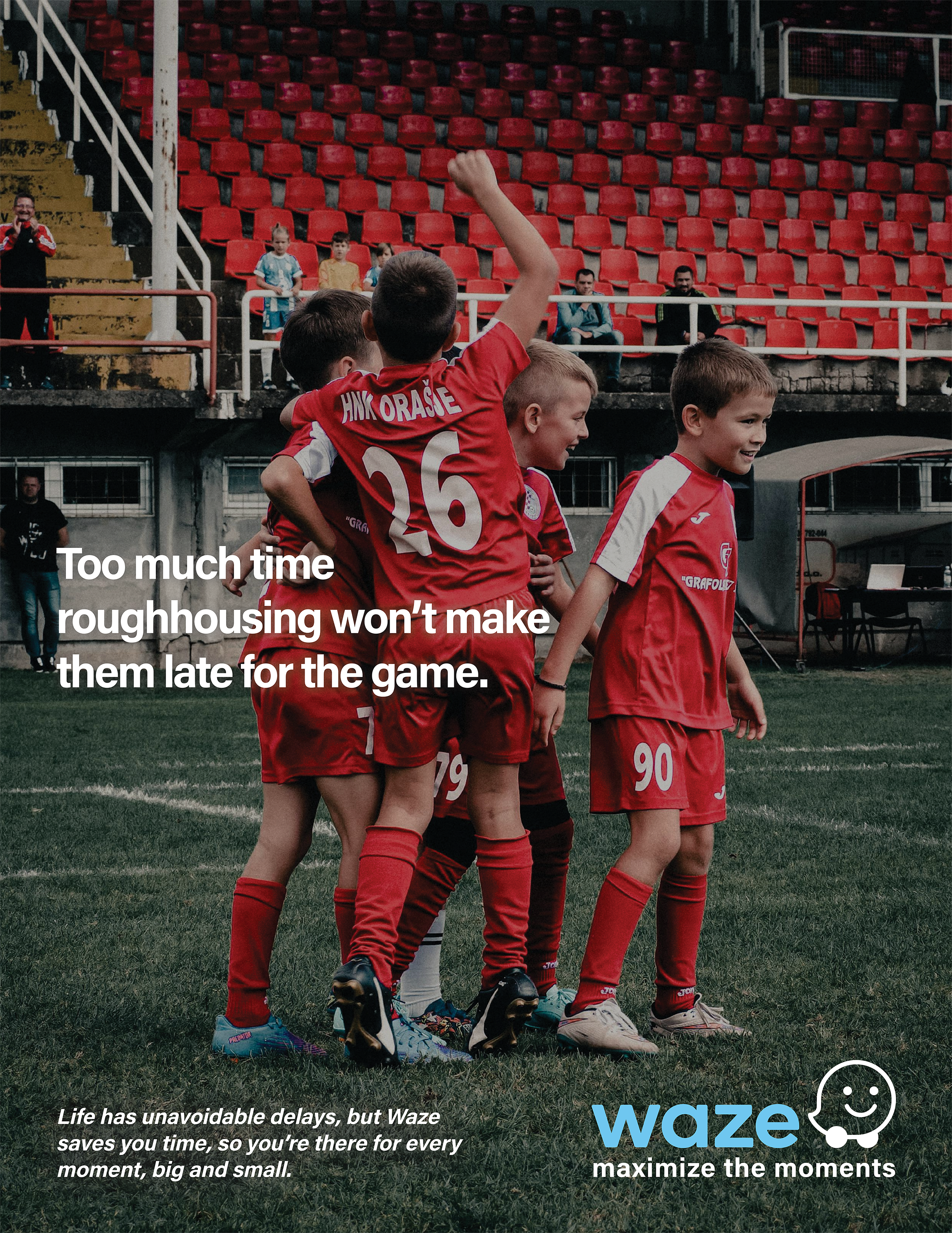 A group of young boys wearing red soccer uniforms with white accents and black cleats standing on a soccer field, with some hugging and others smiling. In the background, there are empty red stadium seats and a few adults, some of whom are sitting an