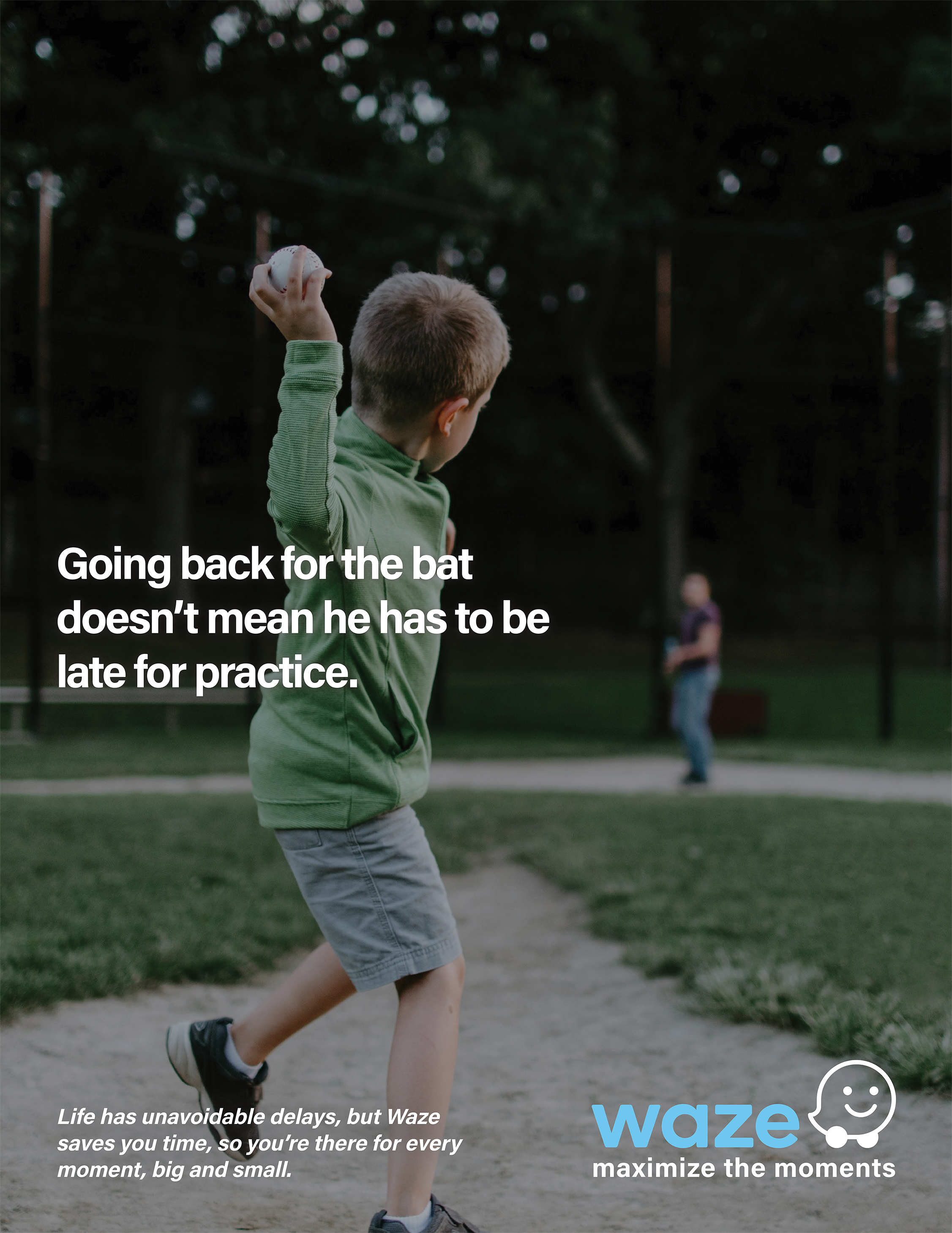 A young boy in a green jacket and shorts is playing baseball in a park. He is holding a baseball in his right hand and is facing away from the camera. There are other children in the background and trees surrounding the area.