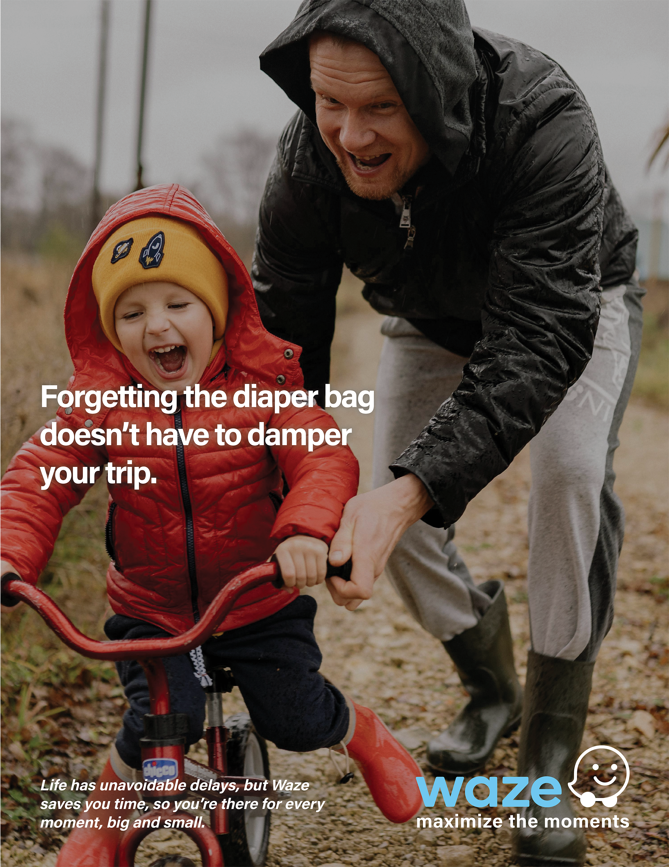 A man and a young child are outdoors, with the man helping the child on a tricycle. The child is wearing a yellow hat and a red puffy jacket, while the man is dressed in a hoodie and black rain jacket. They are smiling and enjoying the moment togethe