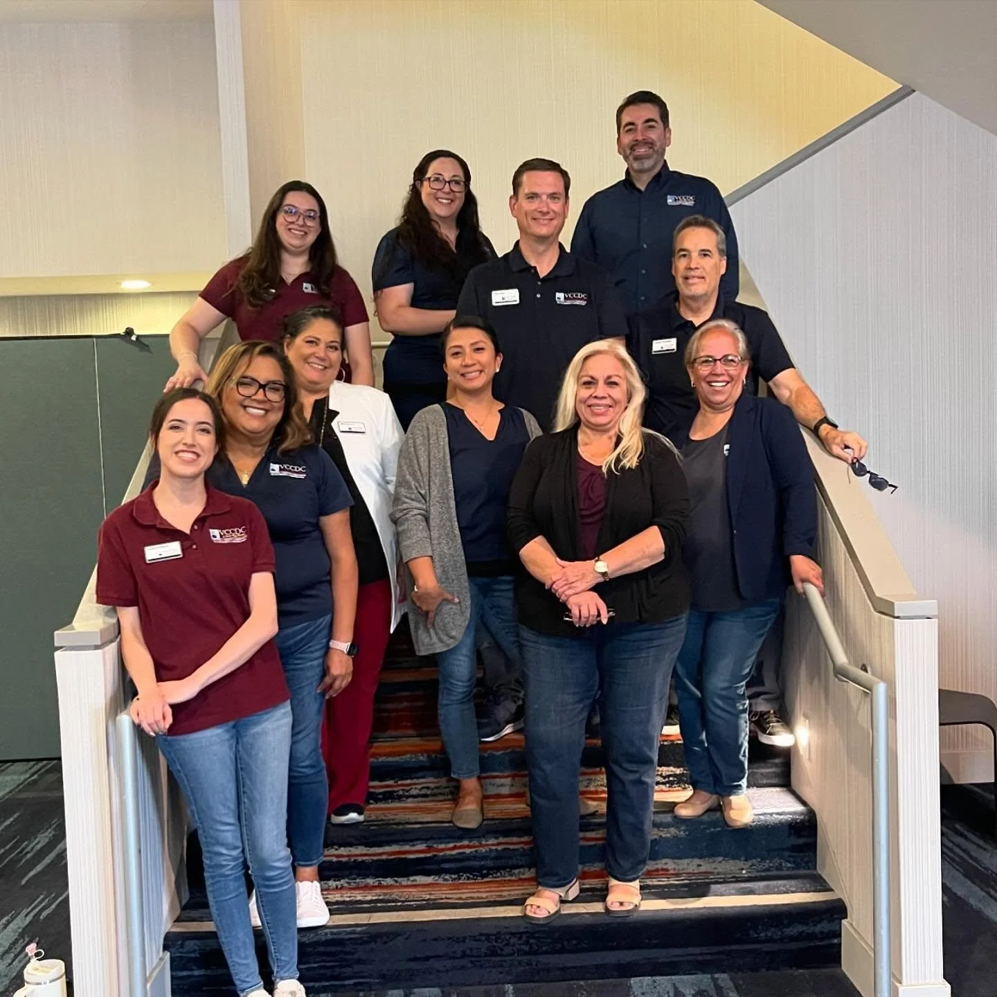 Group of ten people standing on a staircase, smiling, in an indoor setting. Some are wearing corporate polos and name tags, indicating a professional or work-related gathering.