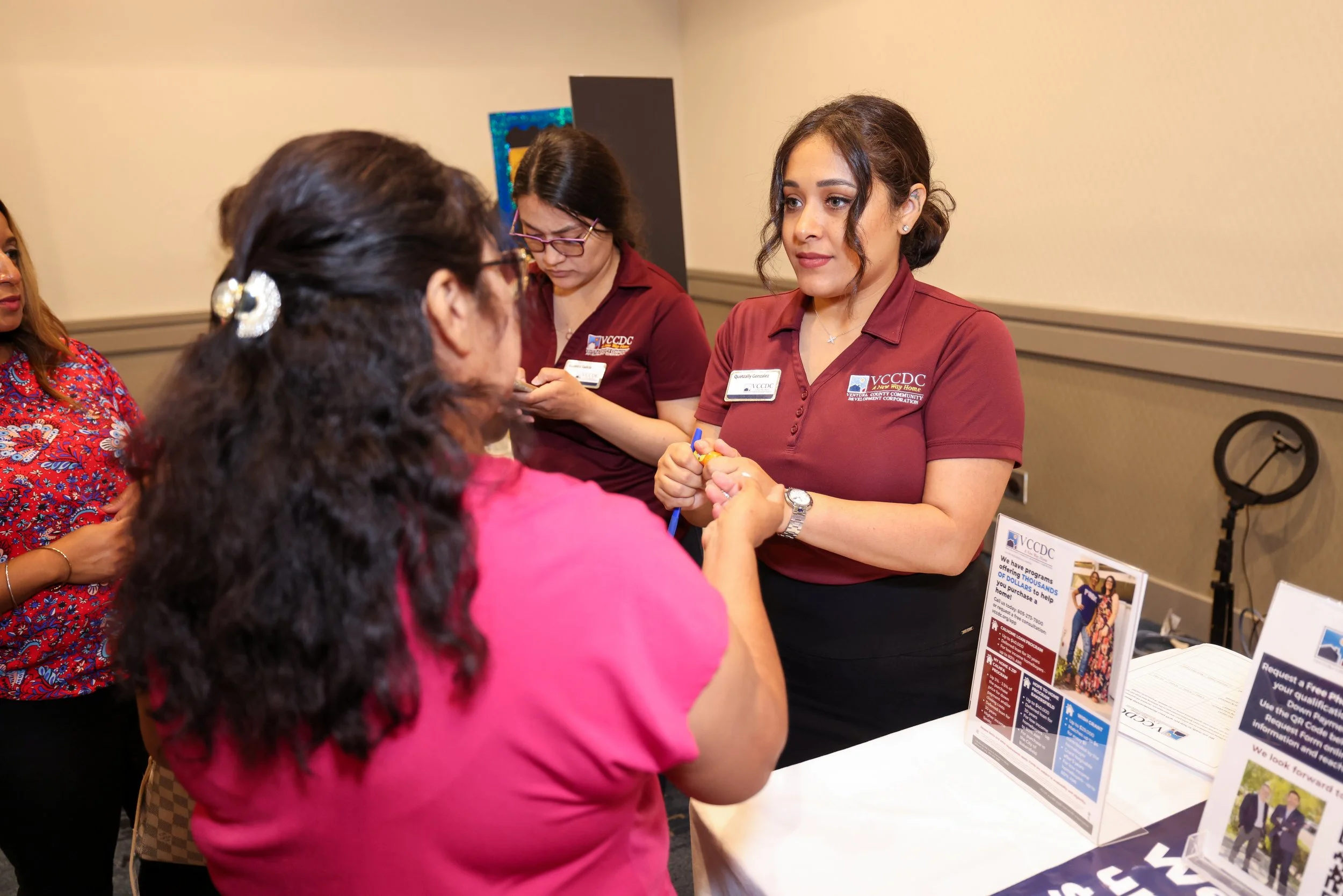 A woman in a pink shirt is receiving information from an event representative at a booth. The booth has pamphlets and signs about a program or event, with two other women in maroon shirts in the background.