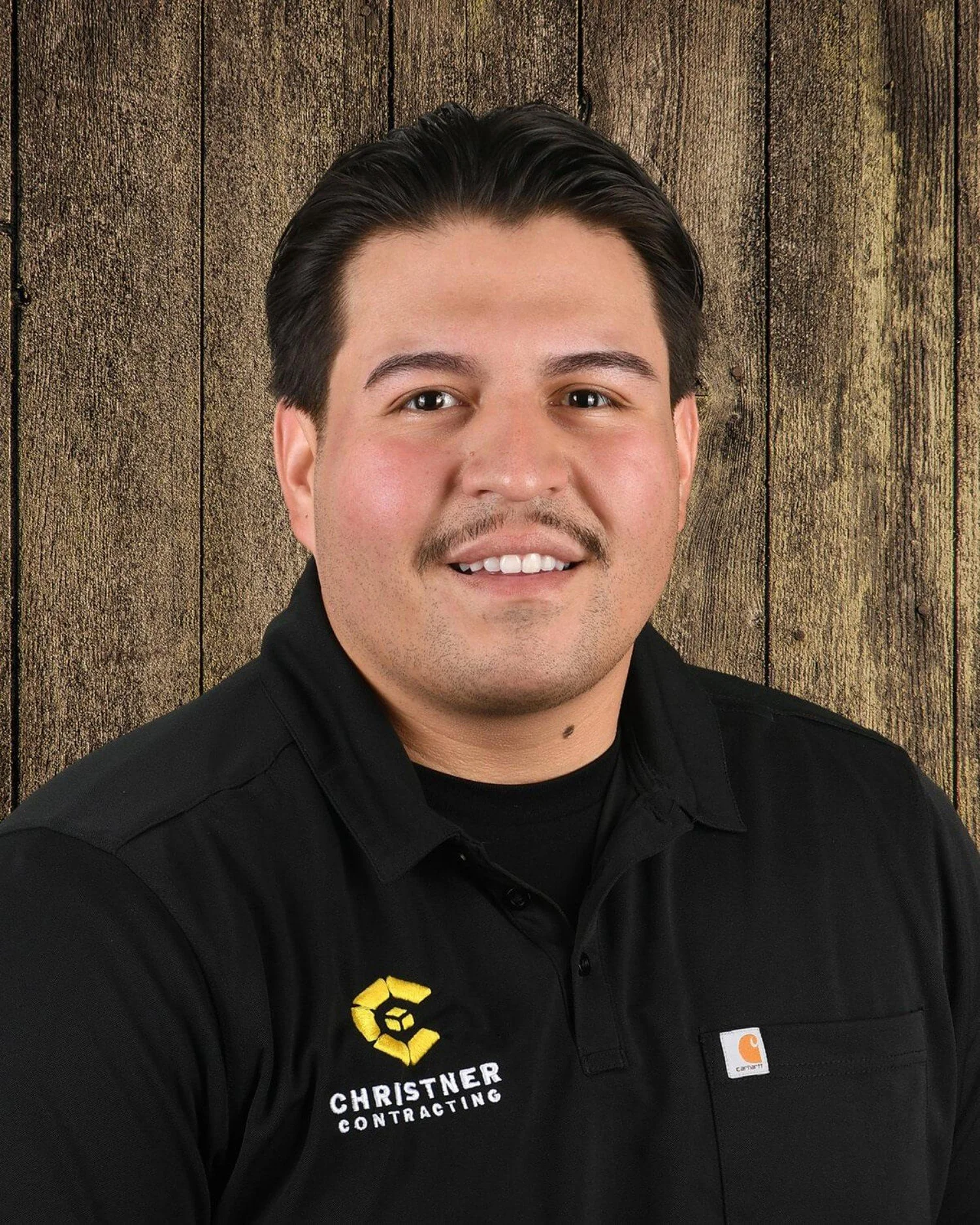 Portrait of a young man with black hair, brown eyes, and a smiling expression, wearing a black collared shirt with a yellow logo on the chest, standing against a wooden background.