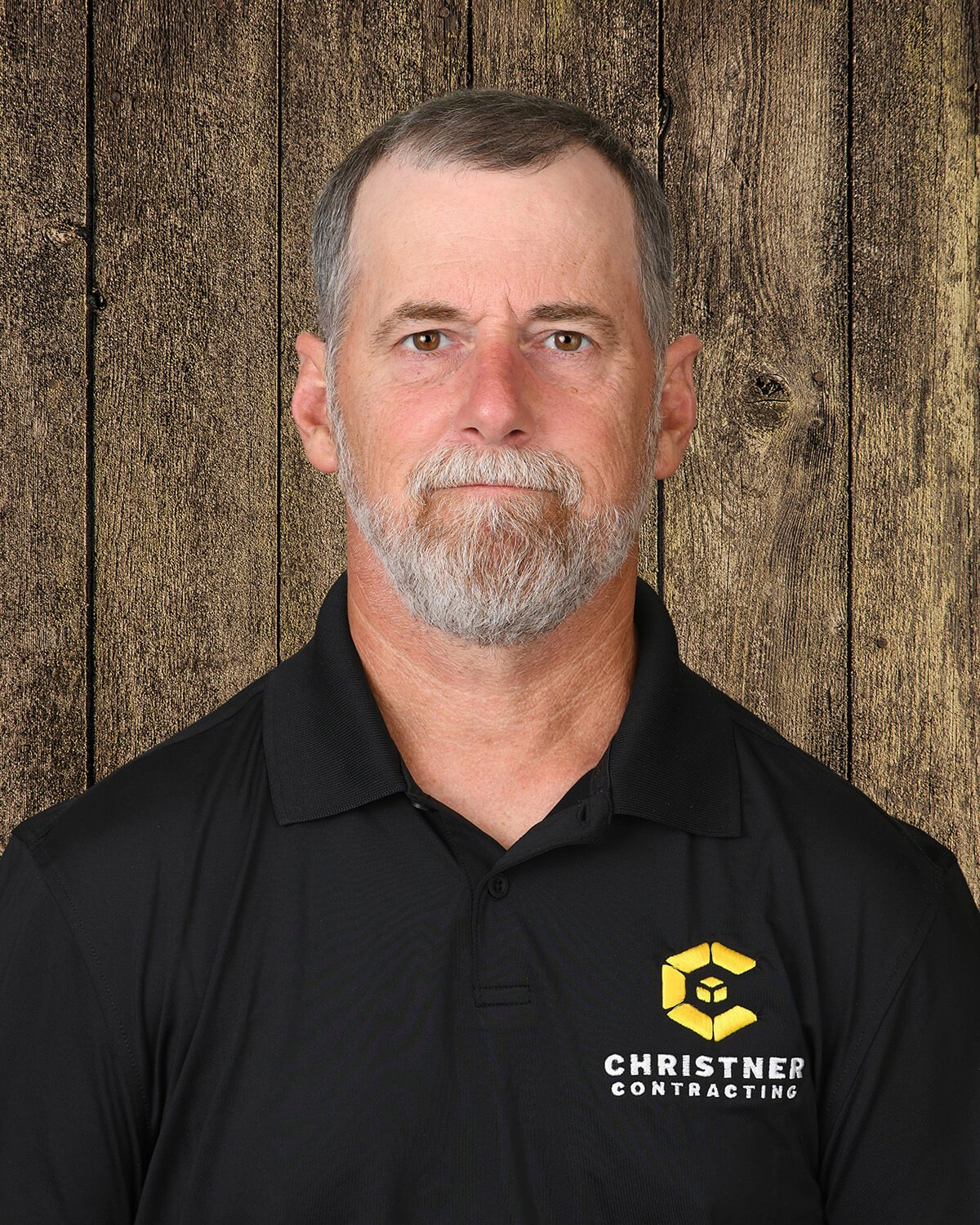 Portrait of a middle-aged man with light brown hair, blue eyes, and a serious expression, wearing a black collared shirt with a yellow logo on the chest, standing against a wooden background