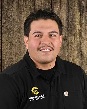 Portrait of a young man with black hair, brown eyes, and a smiling expression, wearing a black collared shirt with a yellow logo on the chest, standing against a wooden background.