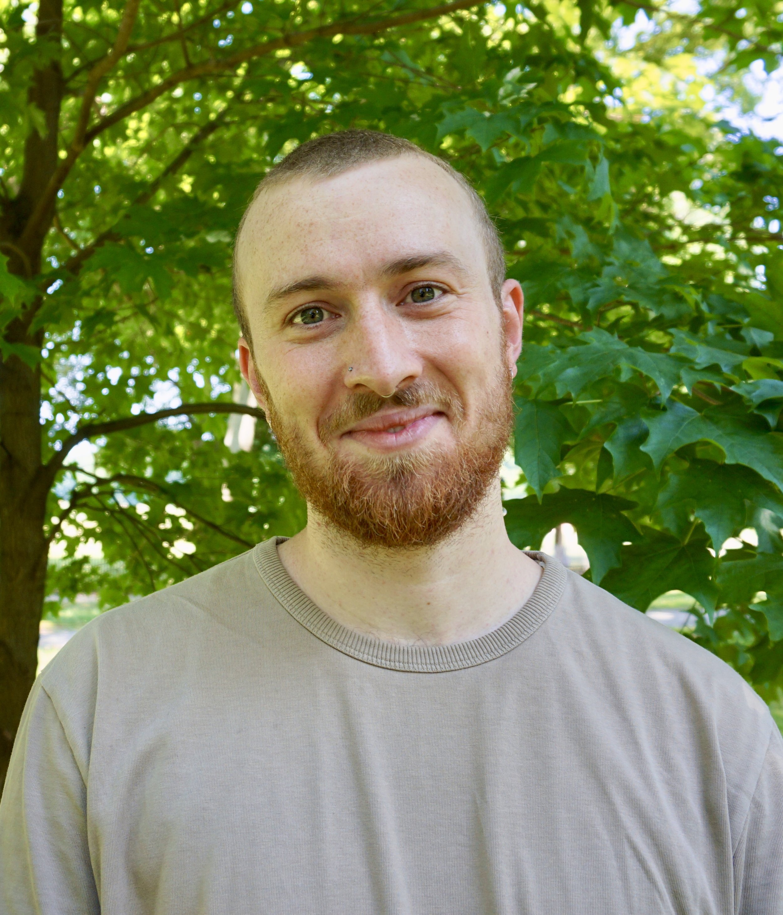 A male therapist with short hair and a beard smiling outdoors with green leafy trees in the background.