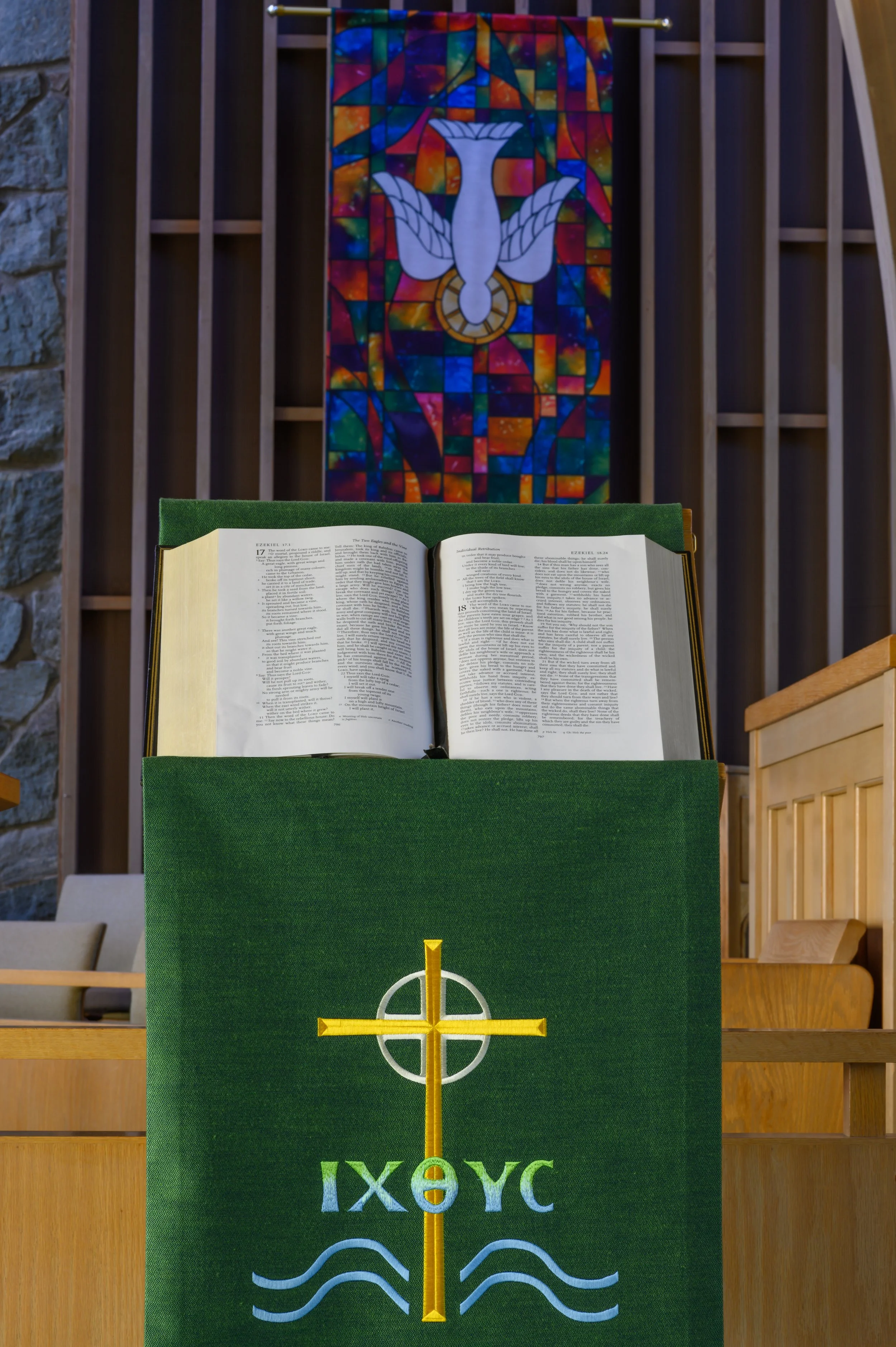 A church altar with an open Bible on a green lectern, decorated with a cross symbol and Greek letters, with a colorful stained glass window featuring a dove in the background.