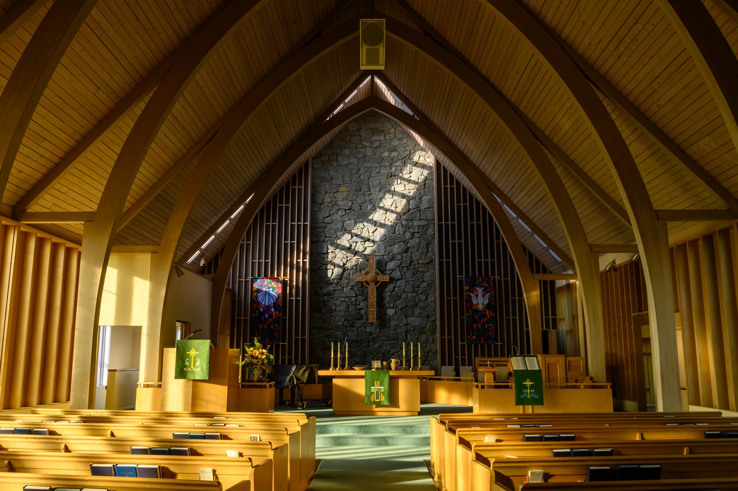 Interior of a church with wooden pews, a stone altar with a cross, candles, flower arrangements, and stained glass windows.