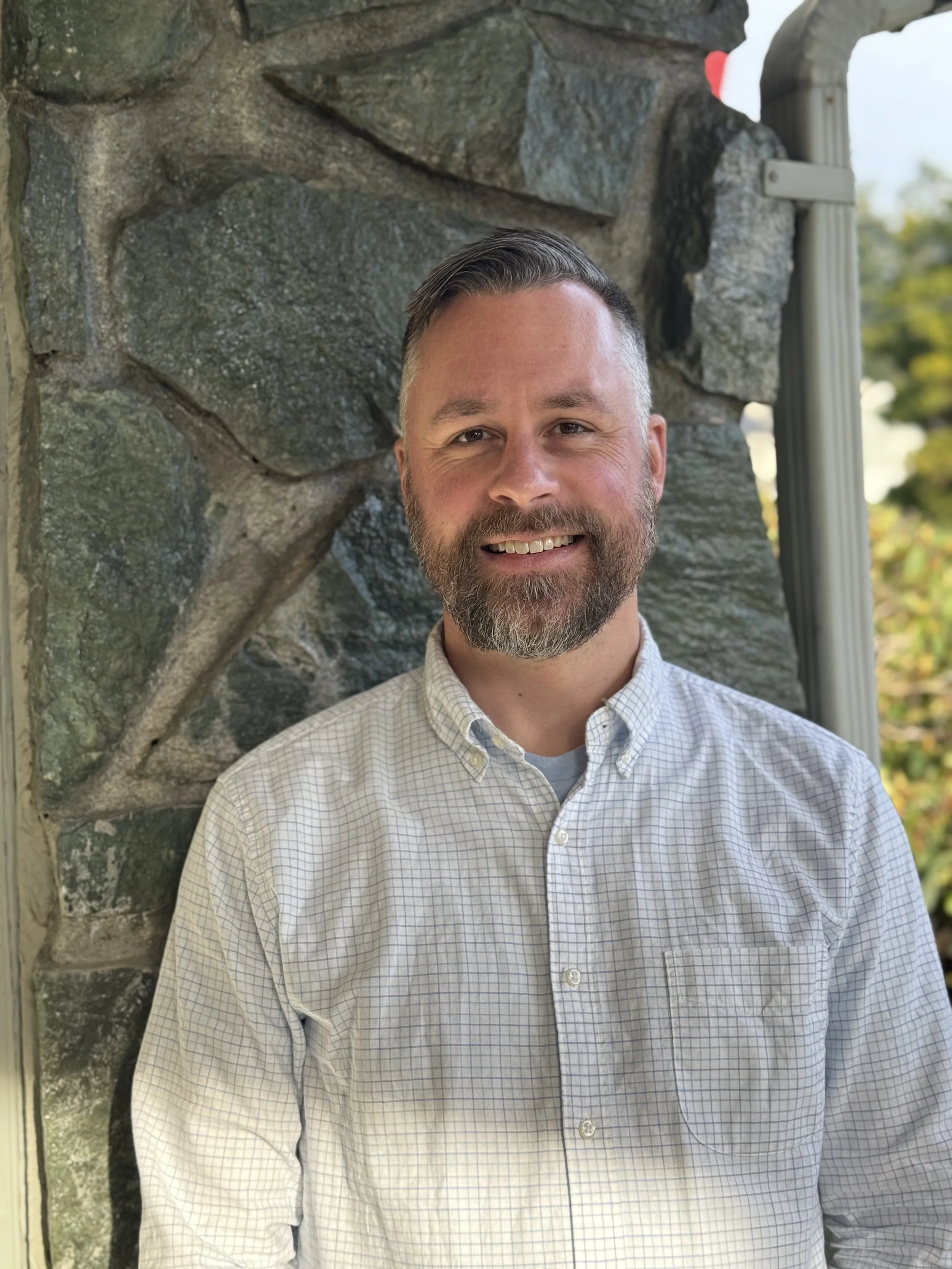 A smiling man with a beard and short hair, wearing a light checked button-up shirt, standing outdoors against a stone wall with trees in the background.