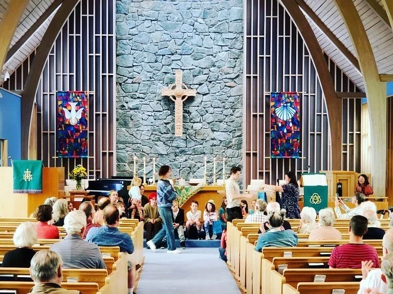 Interior of a church with a congregation, a stone wall, a large wooden cross, stained glass windows, and people seated and standing.