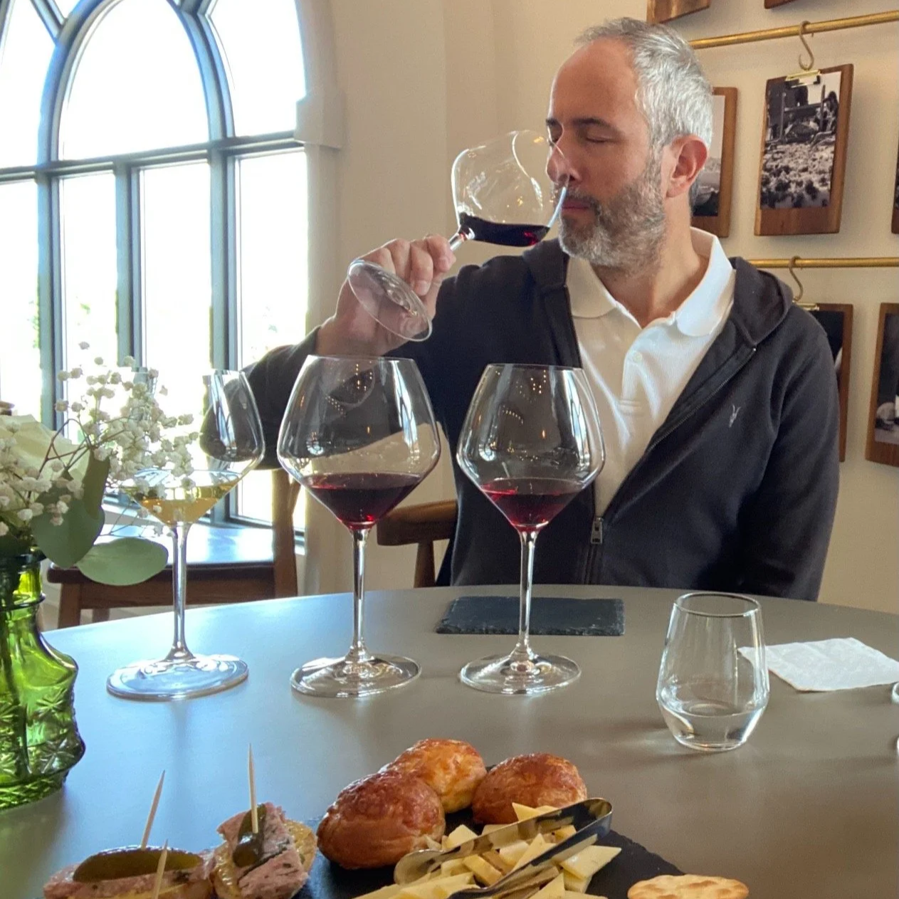 A man with gray hair and beard is tasting red wine from a glass at a dining table with three other glasses of red wine in front of him. The table has a plate of appetizers and a flower arrangement. The setting appears to be a cozy, well-lit restaurant.