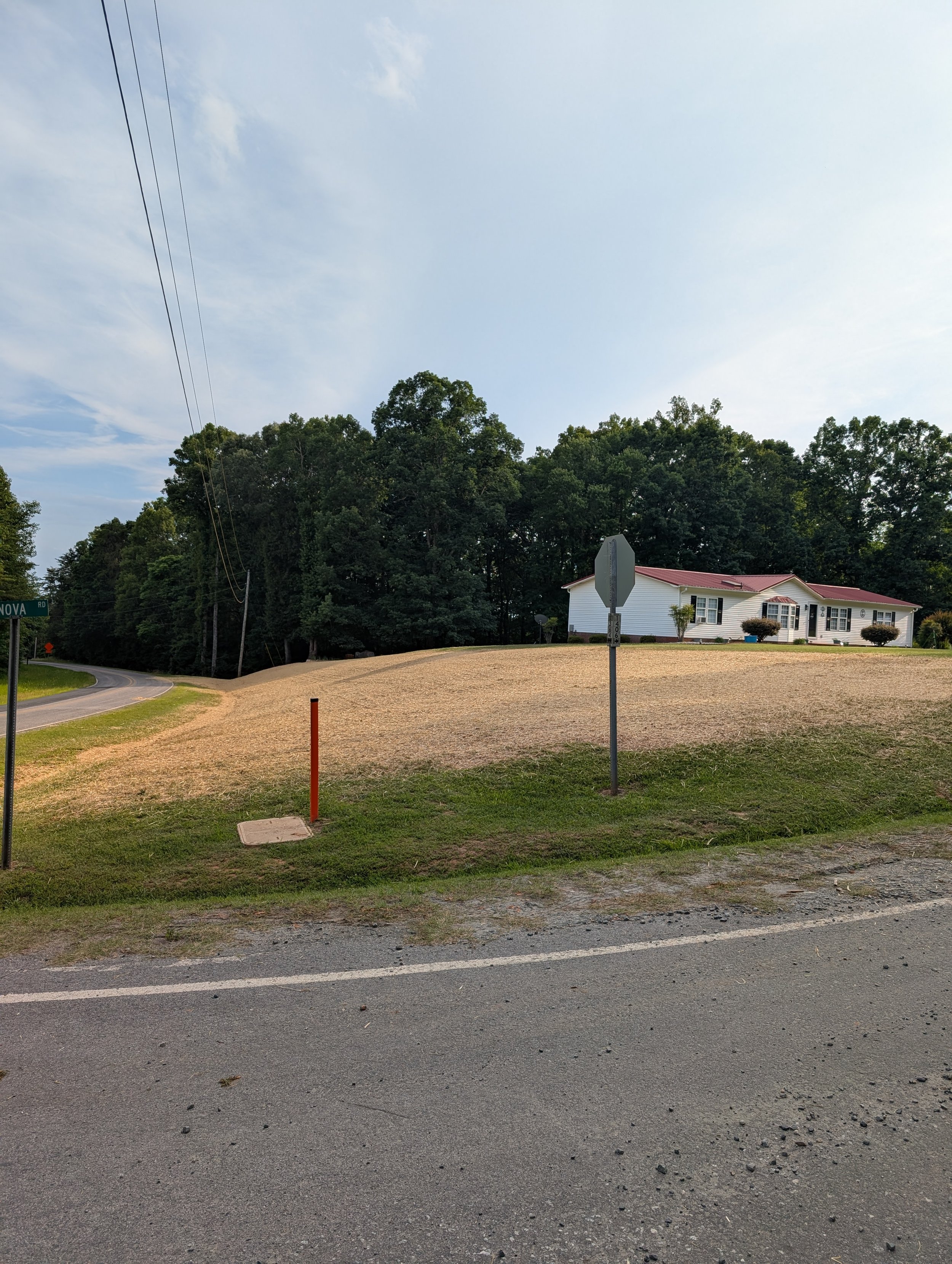 Residential street scene with a white house, trees, power lines, stop sign, and a gravel driveway in a rural area under a partly cloudy sky.