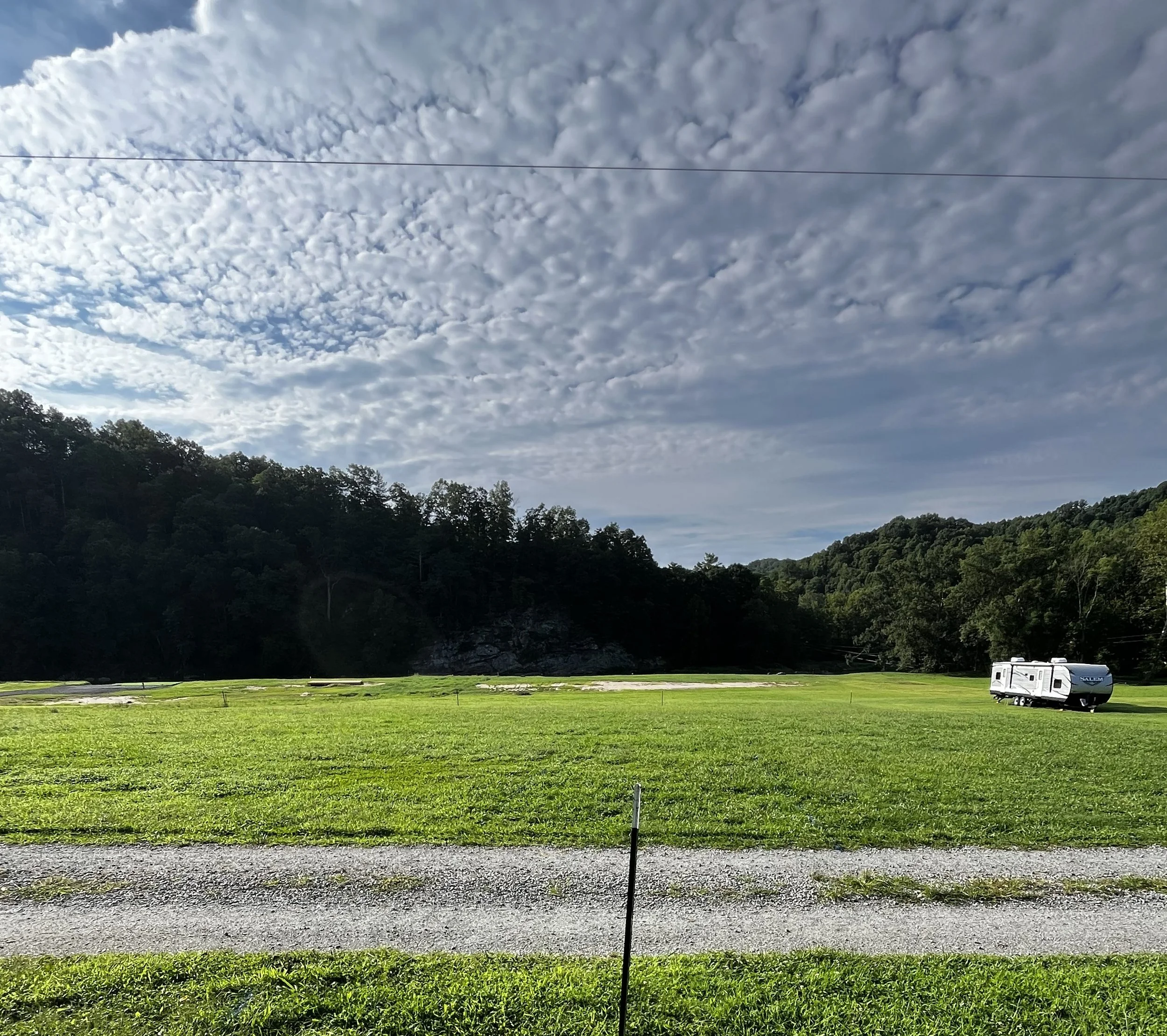A grassy field with trees on a hillside in the background under a partly cloudy sky, and a RV parked in the distance.