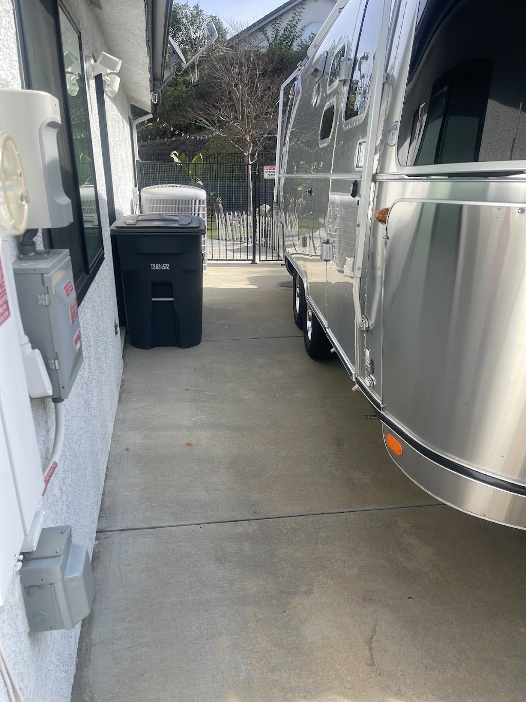 Side of a silver Airstream trailer parked on a driveway next to a house with white walls and windows. A black trash bin and HVAC unit are near the wall. A fence and vegetation are visible in the background.