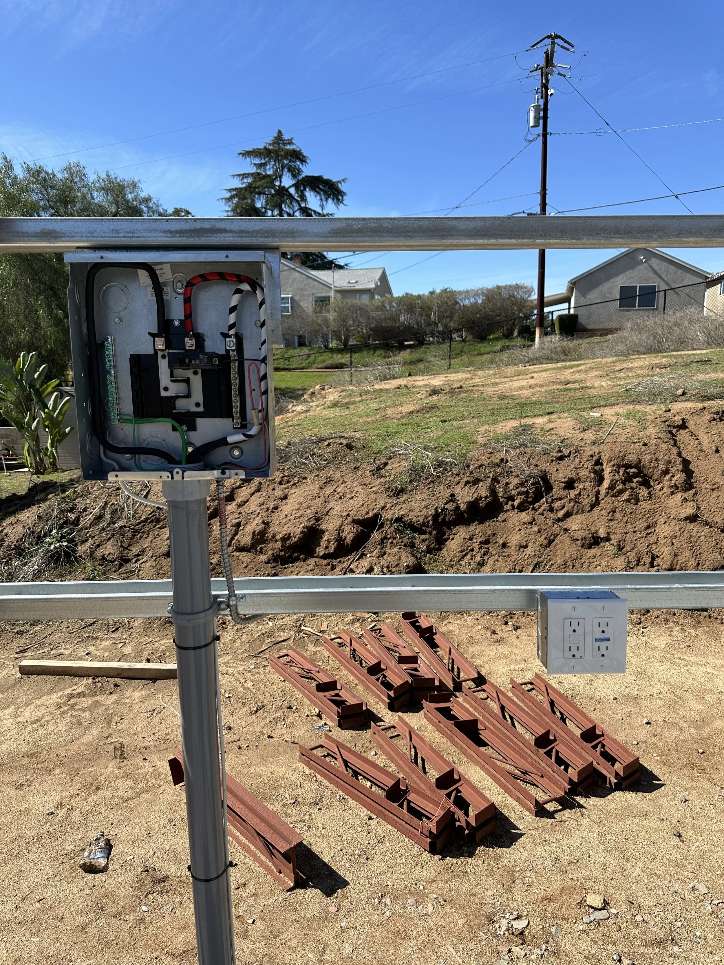 Outdoor electrical junction box with exposed wiring on a pole, partially constructed solar panel mounting brackets on the ground, power outlet, rural background with houses and trees, utility pole with wires.