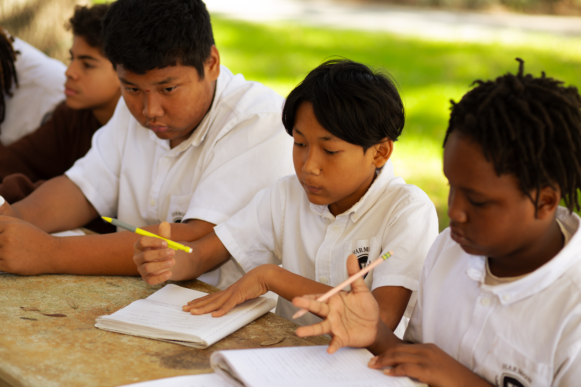 Students studying outdoors at a table, writing in notebooks, wearing white uniforms.