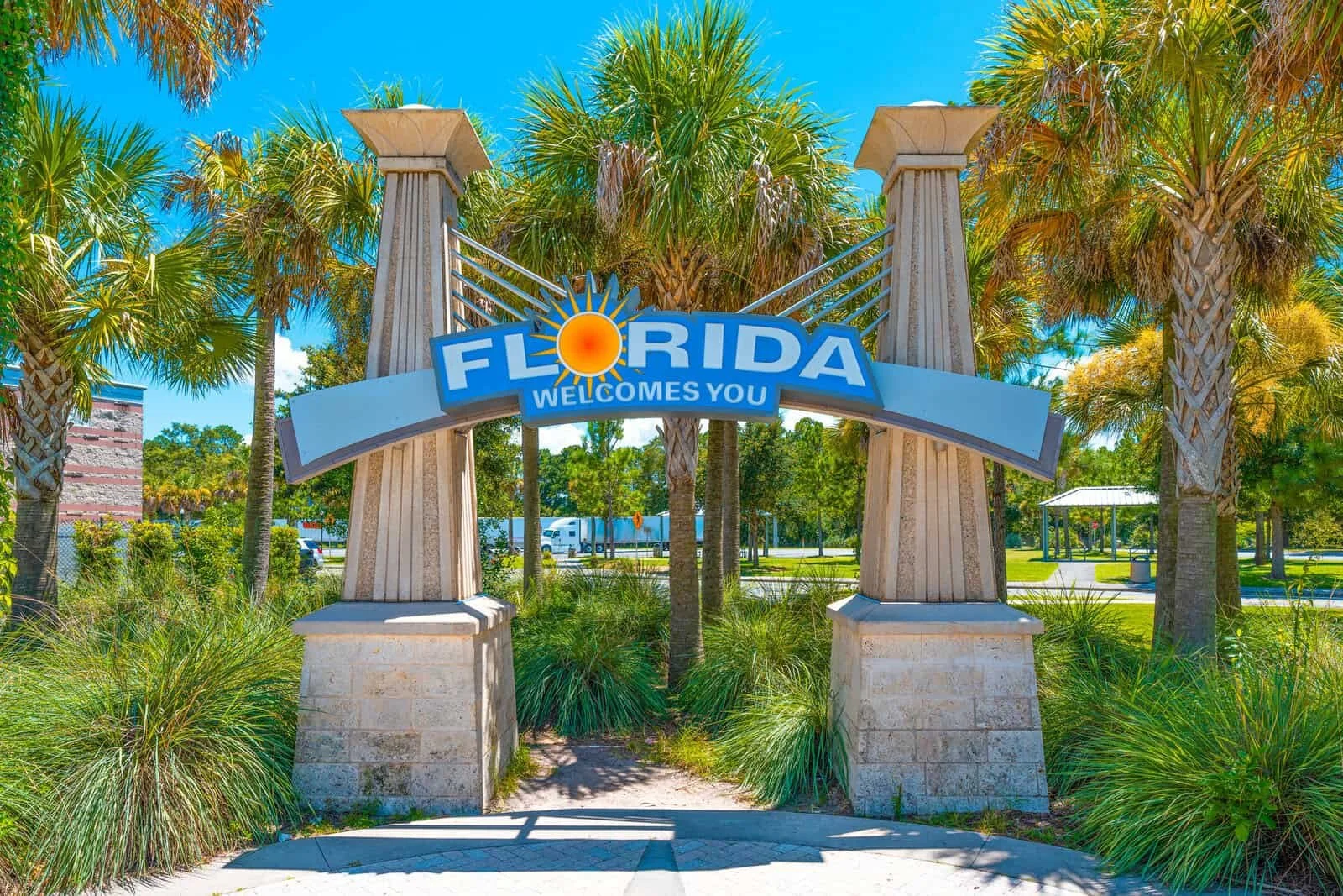 Welcome sign that says 'Florida Welcomes You' with a sunny sun graphic, surrounded by palm trees and greenery, under a bright blue sky.